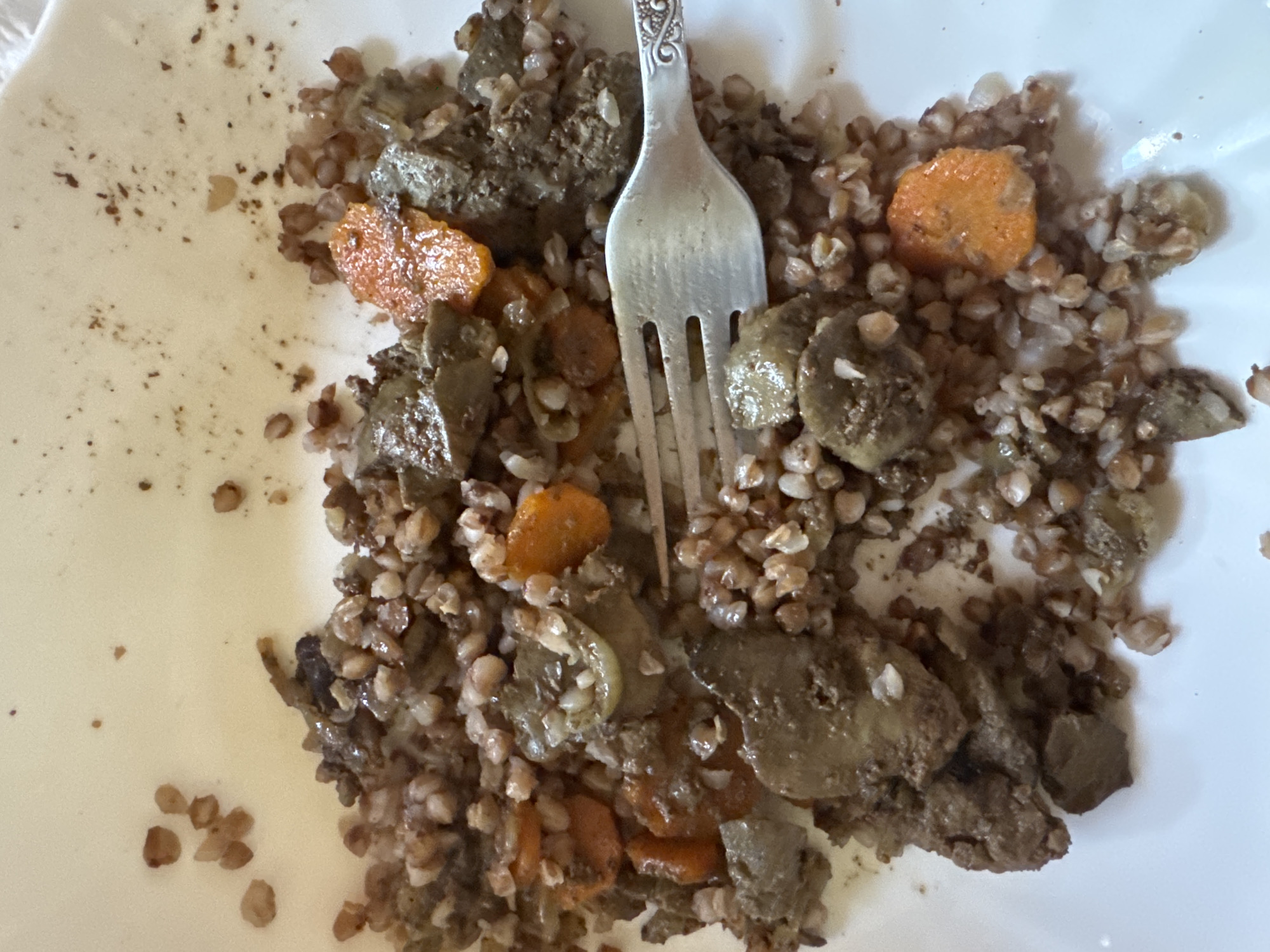 The image displays a close-up, overhead view of a white plate with a portion of buckwheat groats mixed with liver and carrot slices. A silver fork with an ornate handle is partially visible, resting on the food. The food appears to be cooked and served. The background is a plain white surface, likely the plate itself. There are no people or environmental details of a city visible in the image.