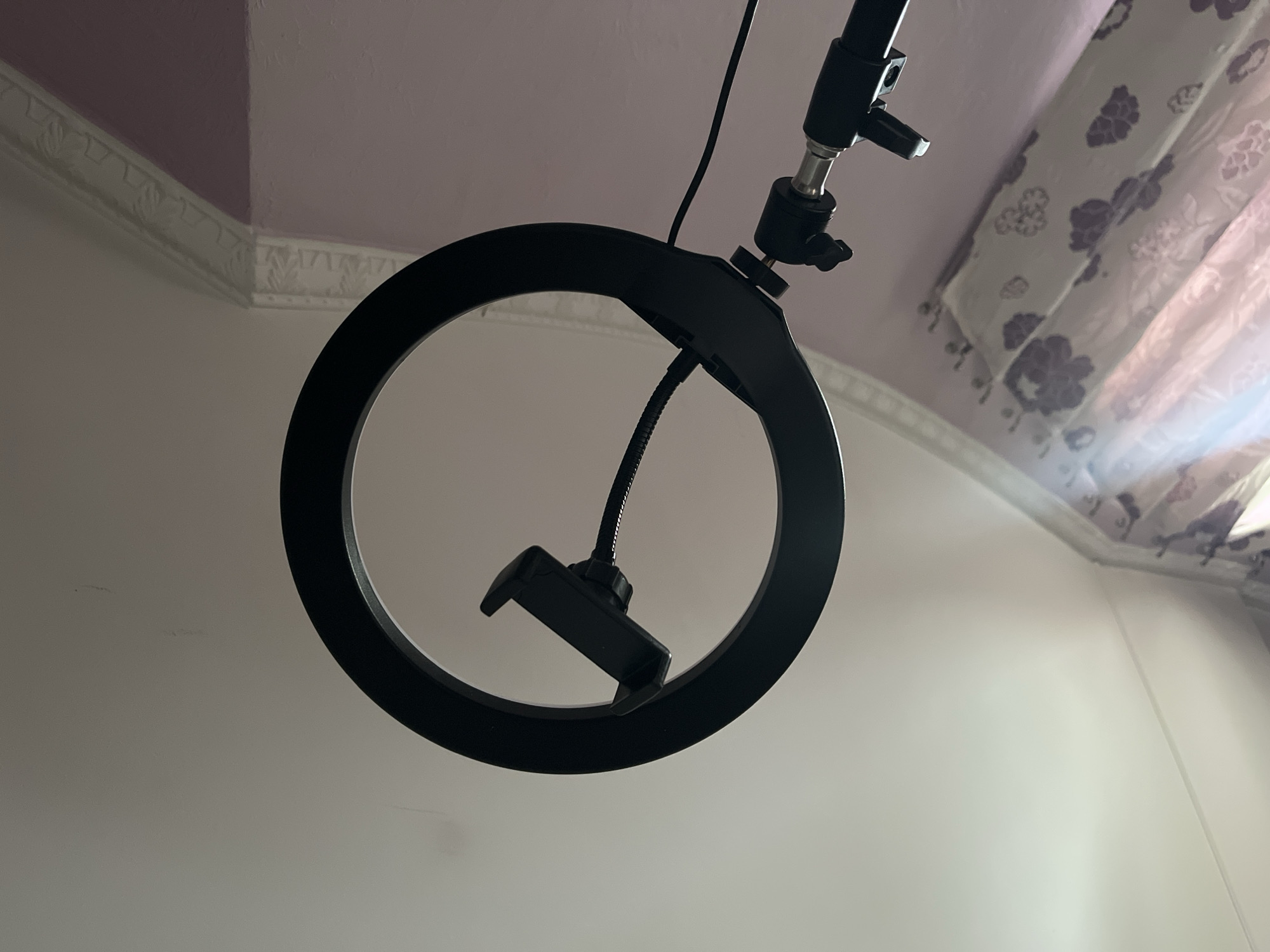 A black ring light with a smartphone holder is mounted on a black tripod. The ring light is positioned in the center of the frame, angled upwards towards a white ceiling. A dark cord extends from the ring light downwards. To the left of the ring light, sheer curtains with a floral pattern are visible, covering a window. The background consists of a light purple wall with decorative molding along the top edge. The scene appears to be indoors, likely in a residential setting, and there are no people or animals present. The image quality is slightly grainy, suggesting it was taken in low light conditions. The location is identified as Tarnivtsi, Ukraine.