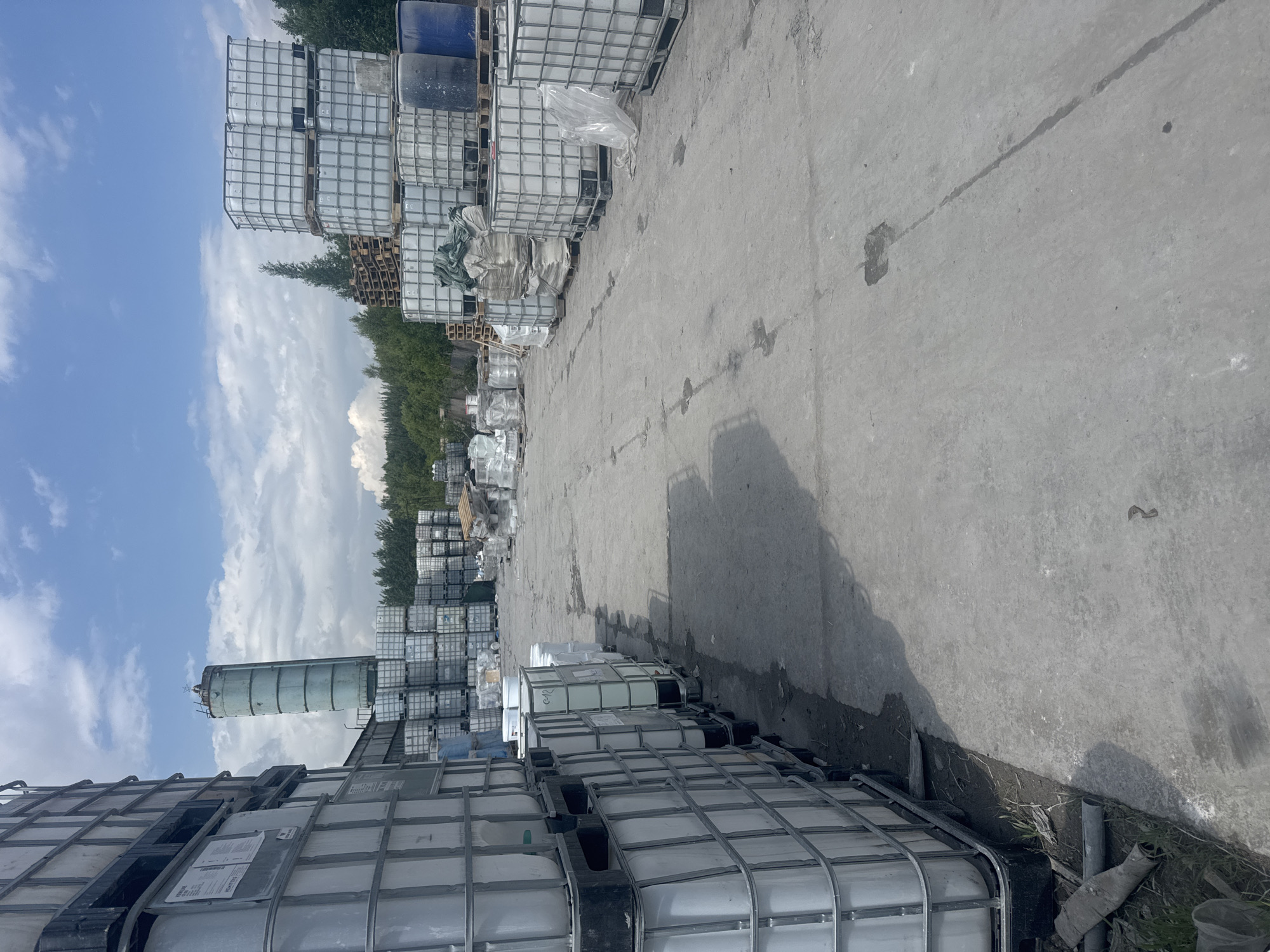 The image depicts an outdoor storage area in the city of Metallostroy, Russia, likely a facility for industrial materials. In the foreground on the left, a tall stack of large white plastic IBC (Intermediate Bulk Container) totes is visible. These totes, encased in a metal cage, are organized in multiple rows and tiers. To the right of the totes, a concrete ground surface extends into the background, with scattered debris and some water or oil stains.

Further into the scene, more IBC totes are arranged in neat stacks, varying in height and configuration. Some are covered with tarps or plastic sheeting, suggesting protection from the elements. In the middle ground, a tall, cylindrical concrete structure, possibly a silo, stands against a bright blue sky with scattered white clouds. Beyond the storage area, a line of trees forms a natural backdrop, with a few wooden pallets also visible amongst the stored materials. The overall impression is one of an industrial yard, orderly and functional, with a variety of large containers holding unspecified materials. No people are visible in the image.
