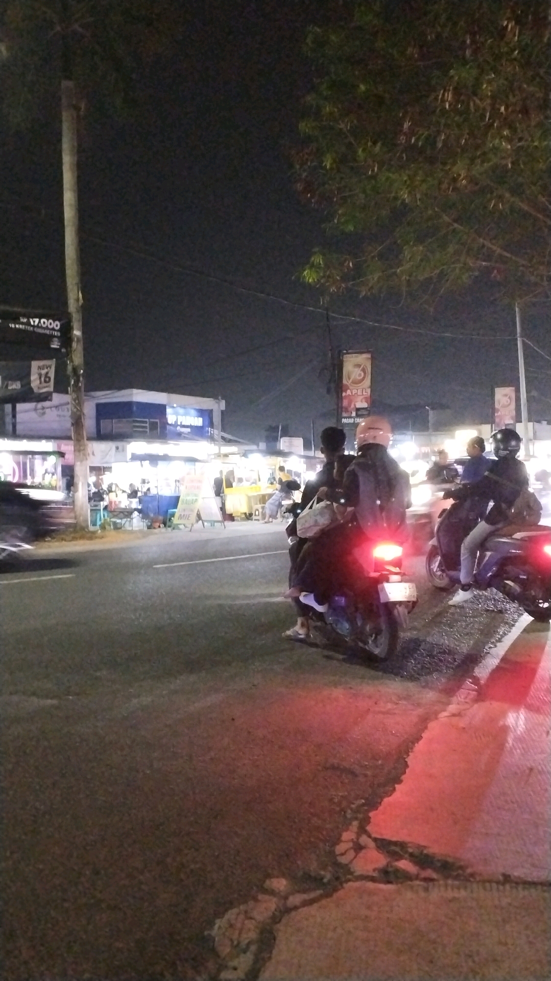 A vertical, night-time, medium-distance shot captures a street scene in Canggu, Indonesia. The foreground is dominated by a dark asphalt road with a distinct white line marking the edge of the lane, which reflects red light from vehicles. The road appears wet or has a reflective surface. In the middle ground, several motorcycles are visible. One motorcycle in the immediate foreground has its red taillight illuminated. This motorcycle carries at least two passengers, one wearing a dark jacket and helmet, and another in the back with a light-colored bag on their lap. Further down the road, other motorcycles are parked or moving, with riders and passengers also visible.
The background reveals a row of brightly lit roadside stalls or small shops. These establishments are illuminated by white and yellow lights, creating a vibrant atmosphere. Signage is present, including a banner with text in Indonesian and a prominent logo featuring the number "76" within a circle. Trees with dark foliage are visible against the night sky, and a utility pole with wires stretches upwards on the left side of the frame. The overall impression is of a busy commercial street at night, likely a popular spot for food vendors or local businesses.