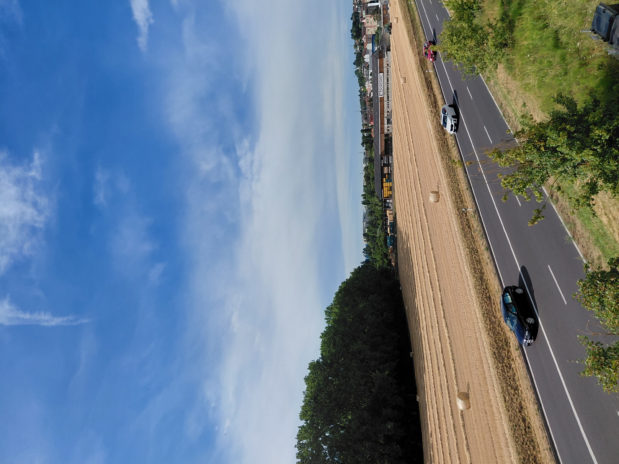 A horizontal shot captures a sunny day in Ecublens, Switzerland, with a clear blue sky dotted with white clouds. A highway runs diagonally across the lower portion of the image. On the highway, a black sedan is in the left lane, and further ahead, a white SUV and a red car are in the right lane. To the left of the highway, a field of harvested wheat is visible, with rows of straw bales scattered across it. A dense line of trees forms the backdrop to the field. In the distance, a town with buildings and a large warehouse is visible on a hill. The overall scene depicts a rural landscape on the outskirts of a city on a bright summer day.