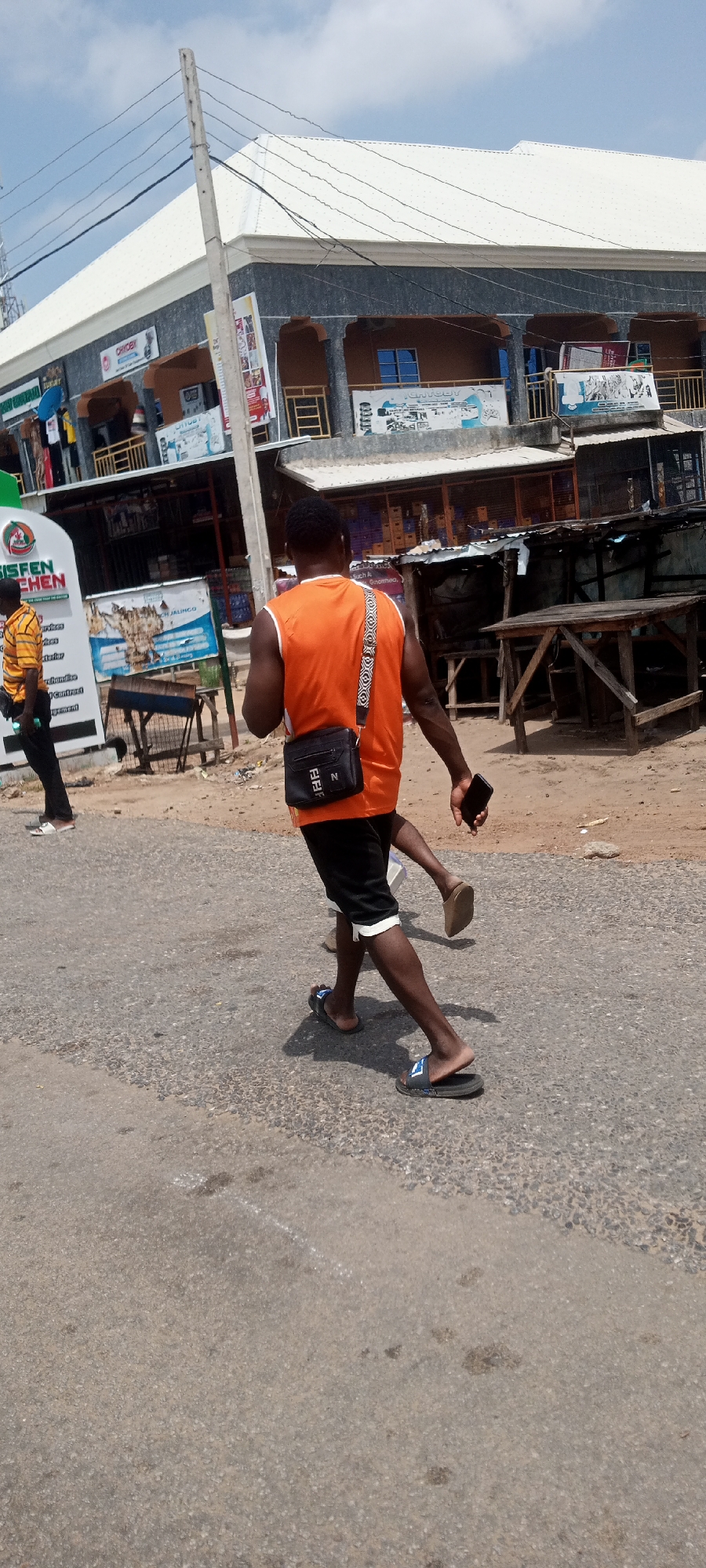 In Jalingo, Nigeria, a man wearing an orange sleeveless shirt and black shorts walks on a paved road, carrying a black shoulder bag and a mobile phone. He is wearing black slides on his feet. To his left, another person wearing a striped orange and yellow shirt and black pants walks in the same direction. Behind them, a two-story building with a white roof and grey exterior stands prominently, featuring multiple shops and blue-tinted windows. Various signs are visible on the building, some with text and images, including one that reads "EISFEN KITCHEN". The environment appears to be an outdoor urban setting with some wooden stalls and tables on the side of the road. Overhead, power lines are strung across the scene. The road surface is asphalt with visible gravel.