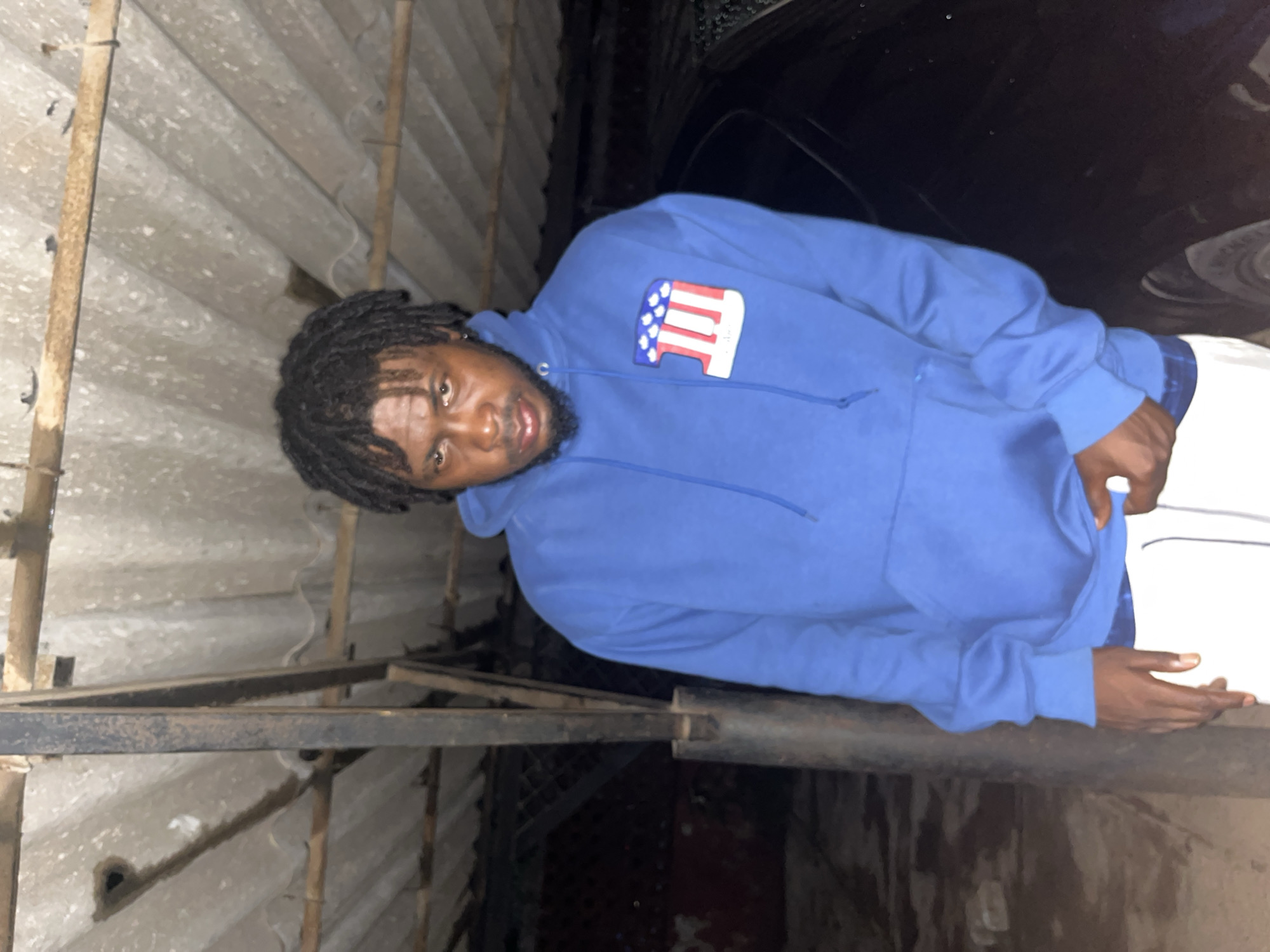 In the city of Jos, Nigeria, a young man with dark, dreadlocked hair stands under a corrugated iron roof supported by rough, wooden beams and a metal pole. He is wearing a bright blue hoodie with a distinctive logo on the left breast featuring red and white stripes and blue stars within a white outline, resembling an American flag motif. His facial expression is serious, with direct eye contact. He is dressed in white sweatpants. Behind him, a dark-colored car is partially visible, suggesting an outdoor setting, possibly a parking area or a sheltered outdoor space. The ground appears to be rough and uneven. The lighting is dim, indicating it might be evening or night.
