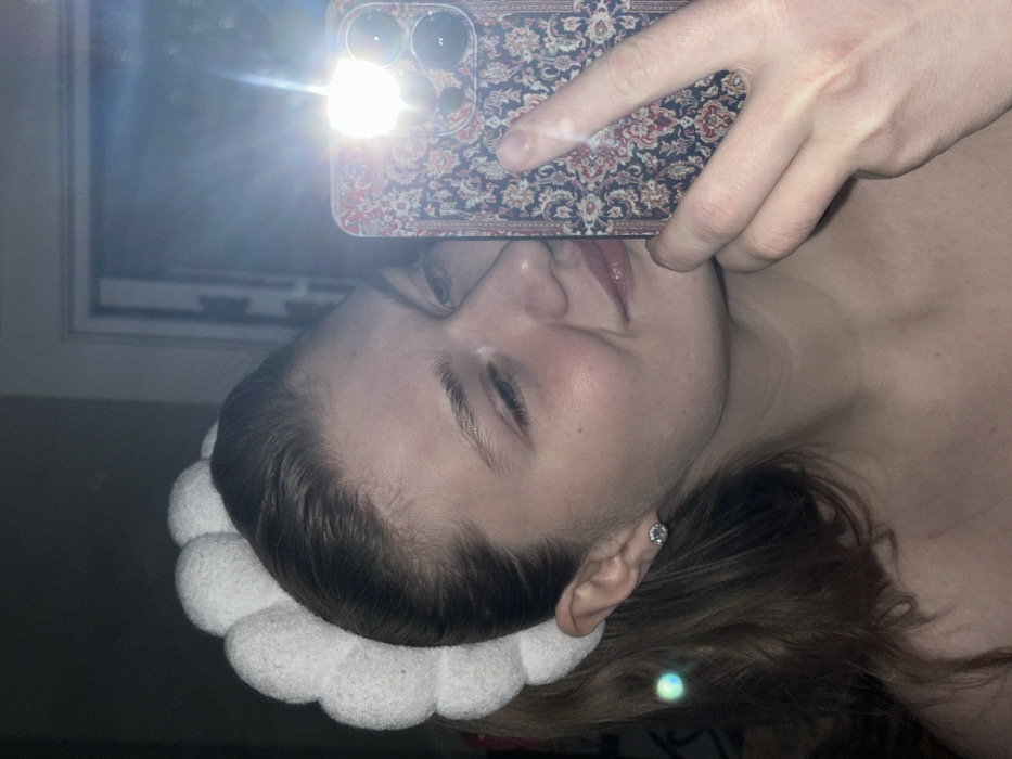 A fair-skinned woman takes a selfie, holding a phone with a floral-patterned case. The phone's flash obscures part of her face. She wears a white, scalloped headband. Her lips are slightly parted, and she wears a subtle gloss. A small, round earring is visible in her ear. The background shows a window and a dark, undefined space. The image is rotated counter-clockwise, presenting a tilted perspective.