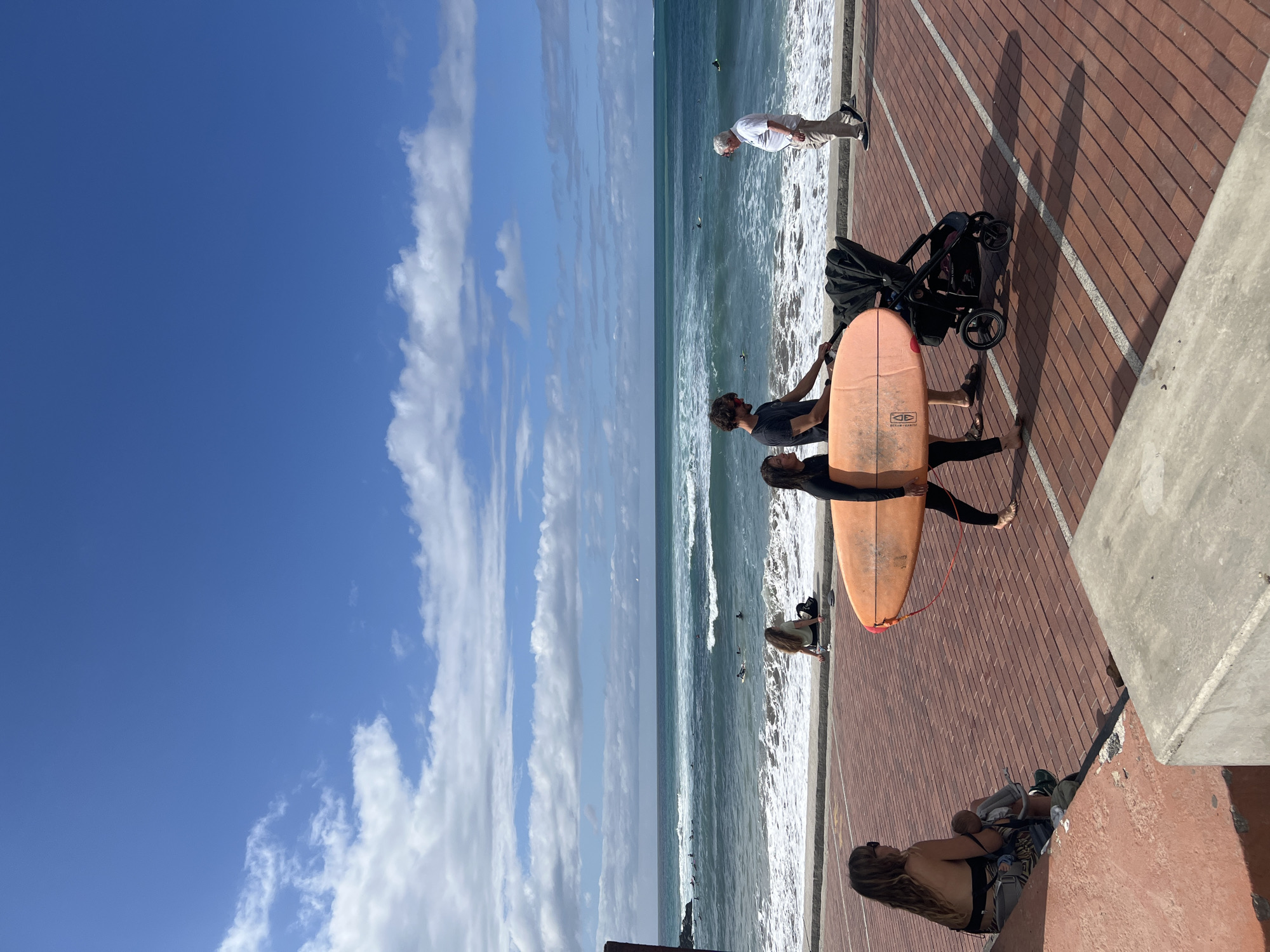 A vibrant, sunny day at a beachfront promenade in Las Palmas De Gran Canaria, Spain. The image captures a lively scene with the deep blue ocean, breaking waves, and a bright sky with scattered white clouds.
In the mid-ground, two individuals stand on a reddish-brown brick promenade, holding an orange surfboard with "DHD" visible on its underside. One wears a dark wetsuit, the other a dark t-shirt. Beside them is a black baby stroller. Further along the promenade to the left, an older man in a white shirt and light trousers stands near the water, observing the ocean. Closer to the water's edge, another person with long, light hair is crouching. Several surfers are visible in the ocean, with some paddling and others riding small waves.
In the foreground, on the right, a person with long, dark, curly hair sits on a concrete ledge, wearing a dark top and patterned shorts, looking towards the ocean. A small child or doll in a carrier rests beside them. Long shadows cast by the people and the stroller on the promenade suggest either morning or late afternoon. The concrete promenade edge runs along the right side of the frame. The overall atmosphere is relaxed and recreational.