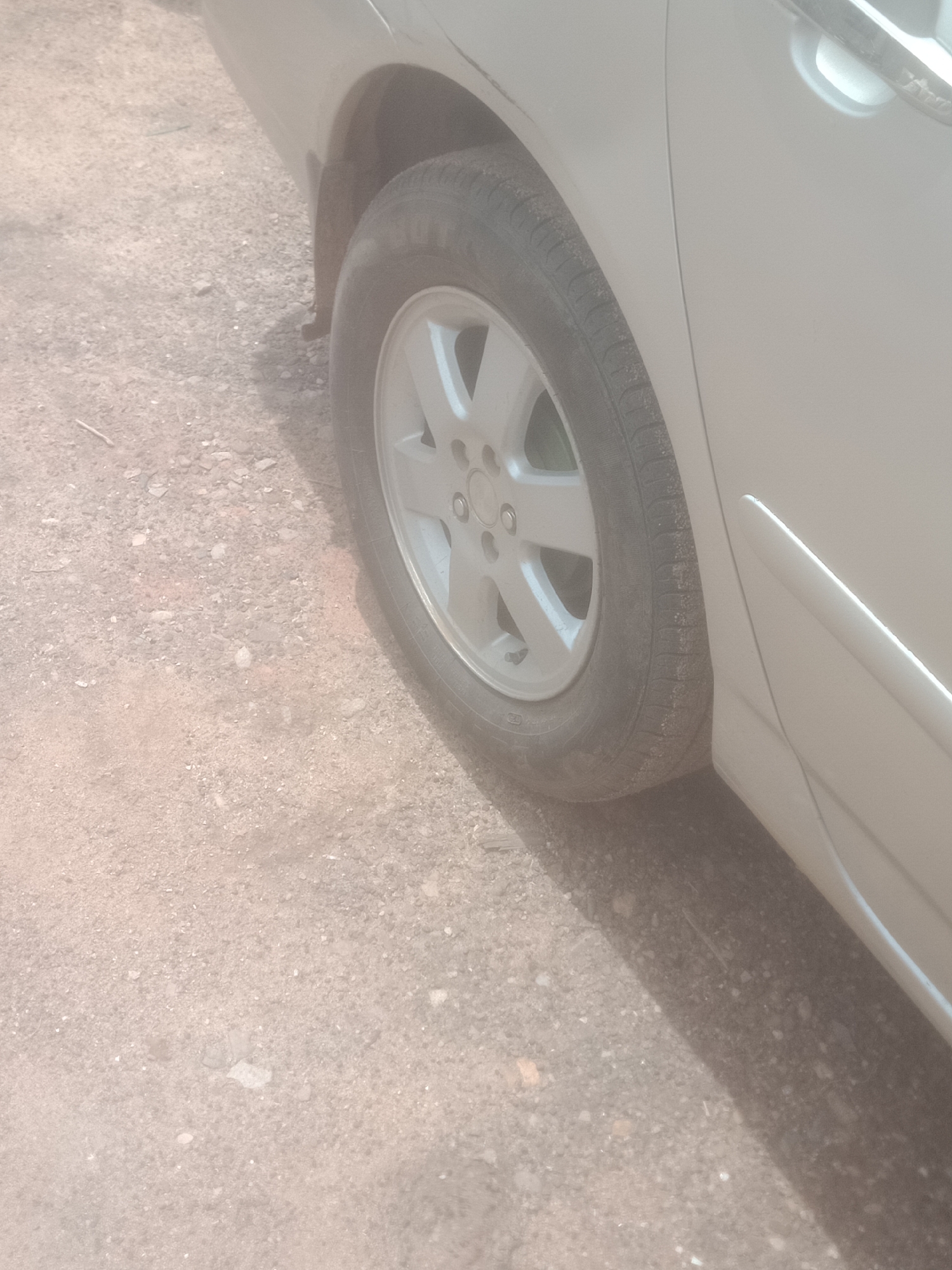 A close-up shot shows the side of a light-colored car, specifically the rear passenger-side wheel and part of the door. The car is parked on a surface of loose gravel and dirt. The tire is dark and appears slightly worn, with some dirt accumulated in the tread. The alloy wheel is silver with a multi-spoke design. The car's paint is a light, neutral color, possibly silver or beige, and it is clean. The door handle is visible, a light-colored rectangular shape. The ground is a mix of small stones, sand, and dirt, with some shadows cast on it, indicating an outdoor environment under daylight. The image is taken from a slightly low angle, focusing on the lower portion of the car.