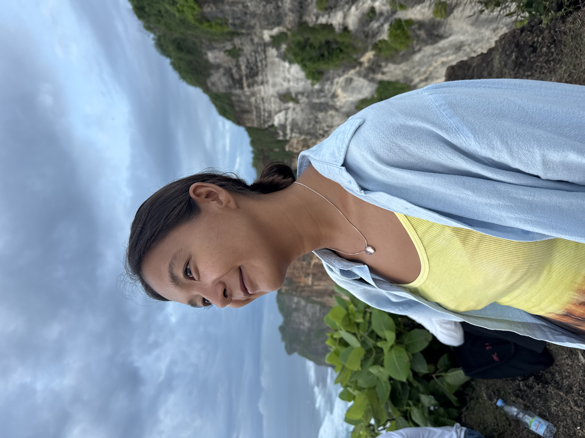 A woman stands on a cliff edge with the sea below, likely at Uluwatu Temple in Kuta Selatan, Indonesia. She wears a light blue shirt over a yellow top and a silver necklace. The background includes a large cliff with green vegetation, a cloudy sky, and ocean waves crashing against the rocks. A few plants are growing in the foreground, near her. Additional items (partially obscured) lie near her including what appears to be a bottle of water, a hat, and a white shirt.