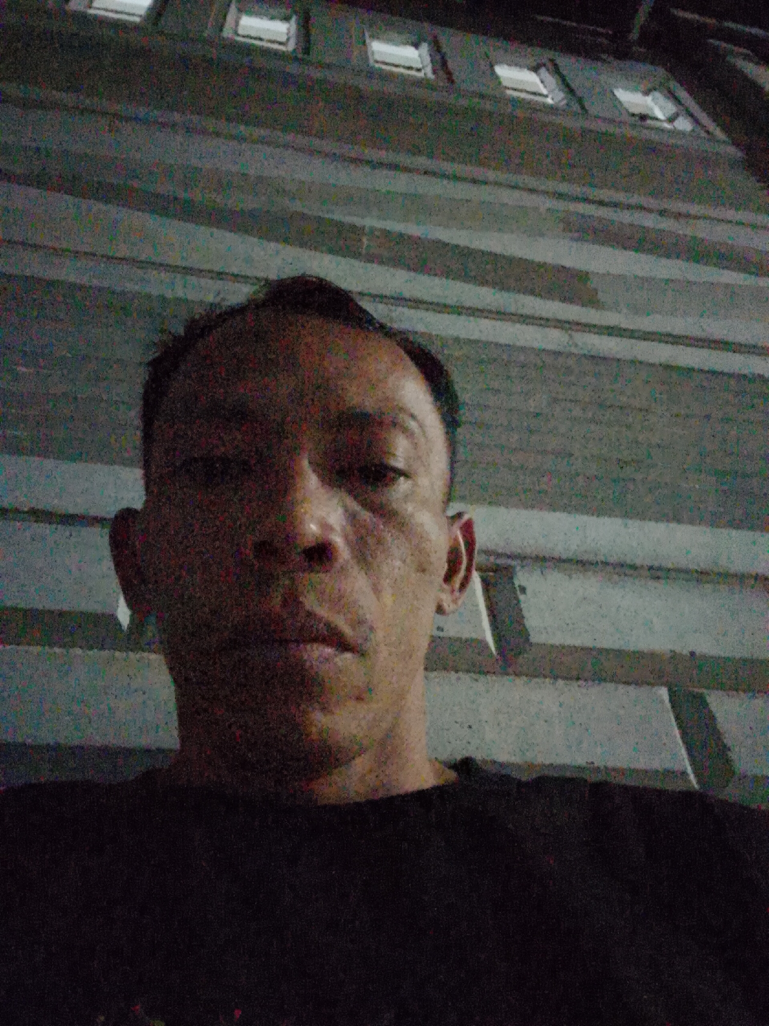 A low-angle, grainy selfie shows a person's head and shoulders against a dark background, with a building facade visible above them. The person, of Southeast Asian descent, has short dark hair and is wearing a dark t-shirt. Their face is in focus, with visible pores and a slightly asymmetrical appearance. The building behind them is grey and appears to have multiple horizontal divisions and evenly spaced rectangular windows. The image was captured in Bukit Ketok, Indonesia.