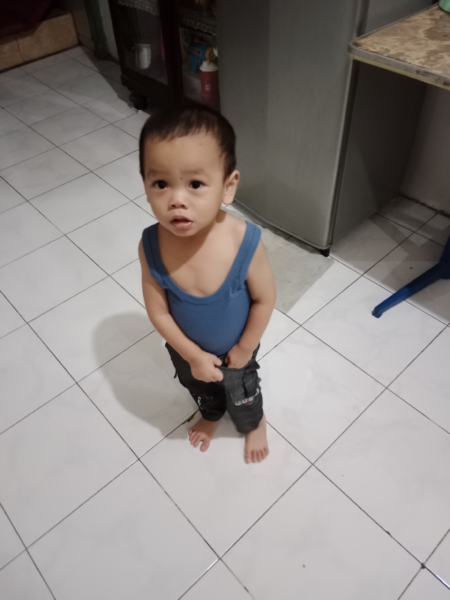 A young boy, appearing to be around two years old, stands on a white tiled floor, looking upwards towards the camera. He is wearing a blue tank top and dark grey pants with pockets. His right hand is inside the front pocket of his pants, and his left hand is resting on his thigh. The boy has dark hair and is looking directly at the camera with an open mouth. The background consists of a tiled floor, a large grey appliance, a dark wooden cabinet with items on shelves, and a blue chair leg. The image appears to have been taken indoors, possibly in a kitchen or living area of a home located in Krian, Indonesia.