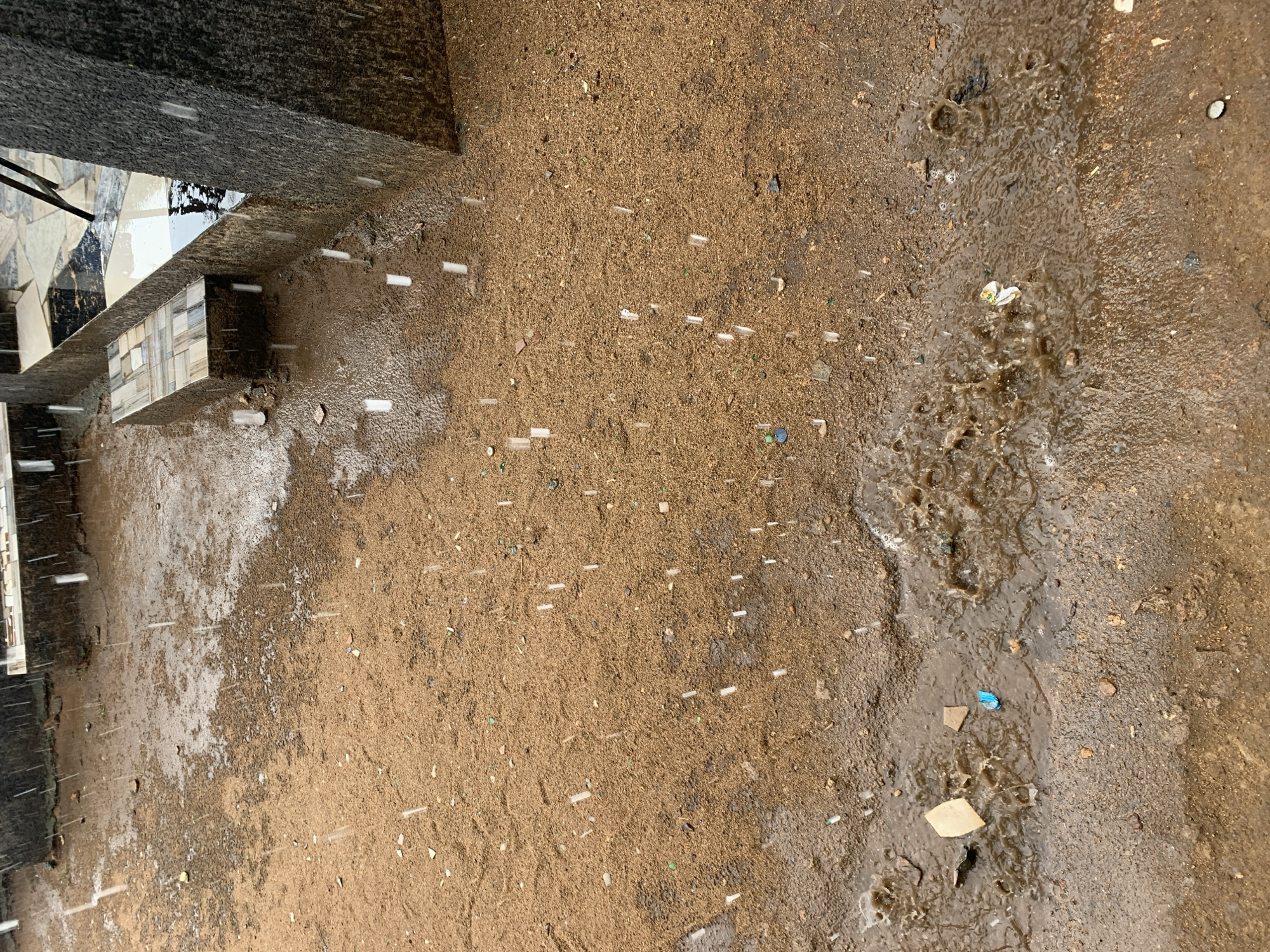 The scene is a ground-level view of what appears to be a dirt or sandy area in New Stadium, Nigeria, likely during or after a rain shower. The ground is primarily brown, with varying degrees of wetness, ranging from damp to muddy. There are numerous small, white, cylindrical objects scattered across the area, resembling short pieces of plastic or other debris. Closer to what looks like a building structure in the upper-left of the picture, the ground is more saturated with water and reflects light. A small stream of water is running across the ground on the right side, creating a muddy area with additional debris. The building structure has a dark stone or concrete base, with what looks like tile details. The environment suggests an outdoor area near a building, possibly a construction site or an undeveloped part of the city.