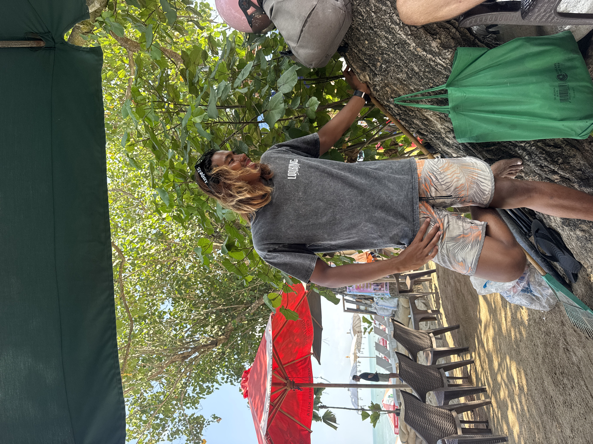 A young man with long, blonde hair and sunglasses is standing on a sandy beach in Kuta, Indonesia, leaning against a large tree. He is wearing a grey t-shirt and patterned shorts. To his right, another person, partially visible, is wearing a grey jacket and a pink helmet. In the background, there are red and white umbrellas, beach chairs, and the ocean. A large green tote bag hangs from the tree. The scene appears to be in a shaded area under a large umbrella.