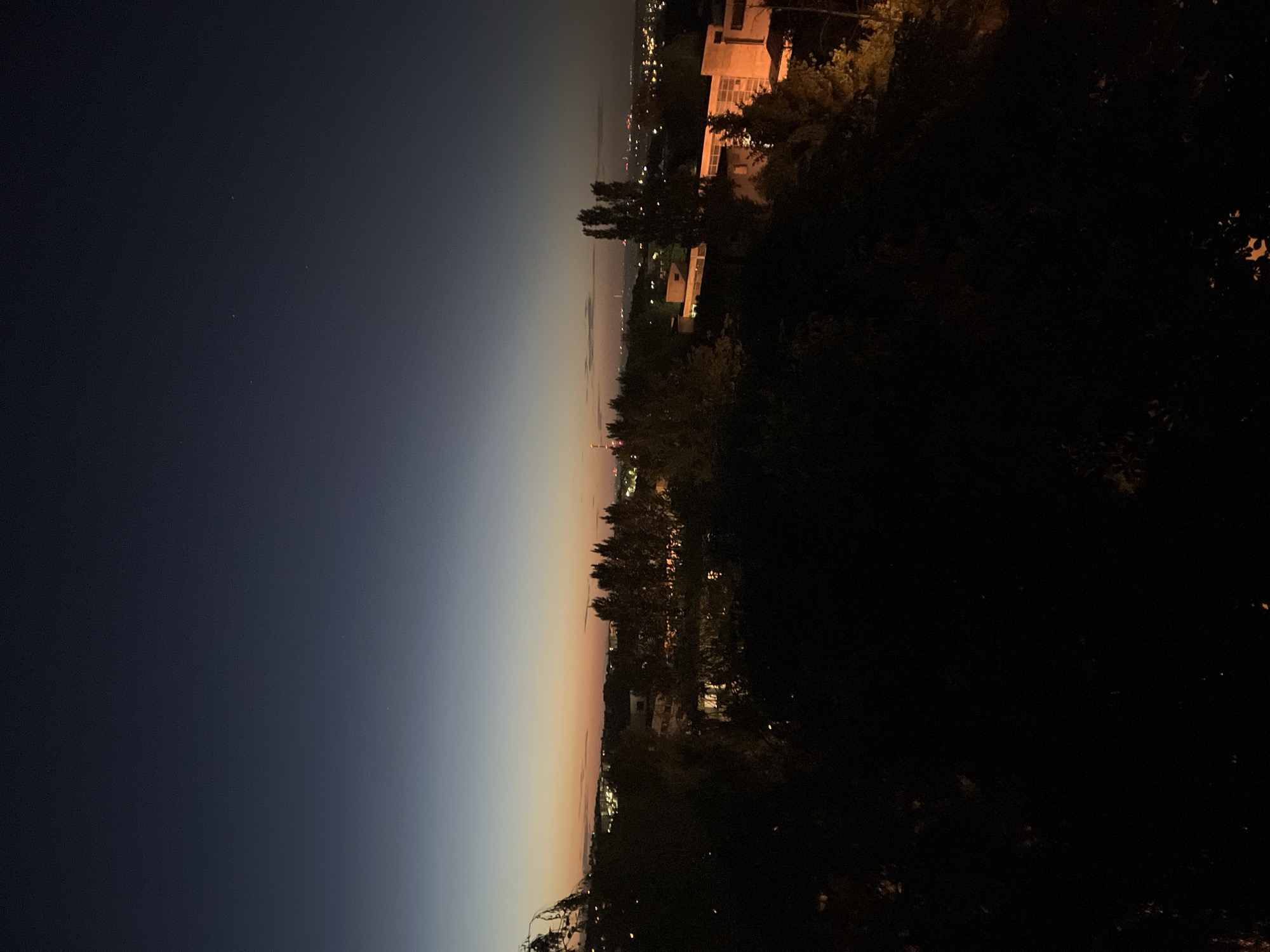 The image captures a night cityscape viewed from an elevated position, potentially a hillside. The sky transitions from a dark blue on the left to an orange and light pink gradient near the horizon. A distant city skyline is visible along the horizon line, punctuated by scattered lights. Buildings are illuminated within the city, and some stand out against the dark sky. In the foreground, dark silhouettes of trees and vegetation occupy the right side of the frame, obscuring the view. The location is implied to be in the city of Unknown, Unknown.