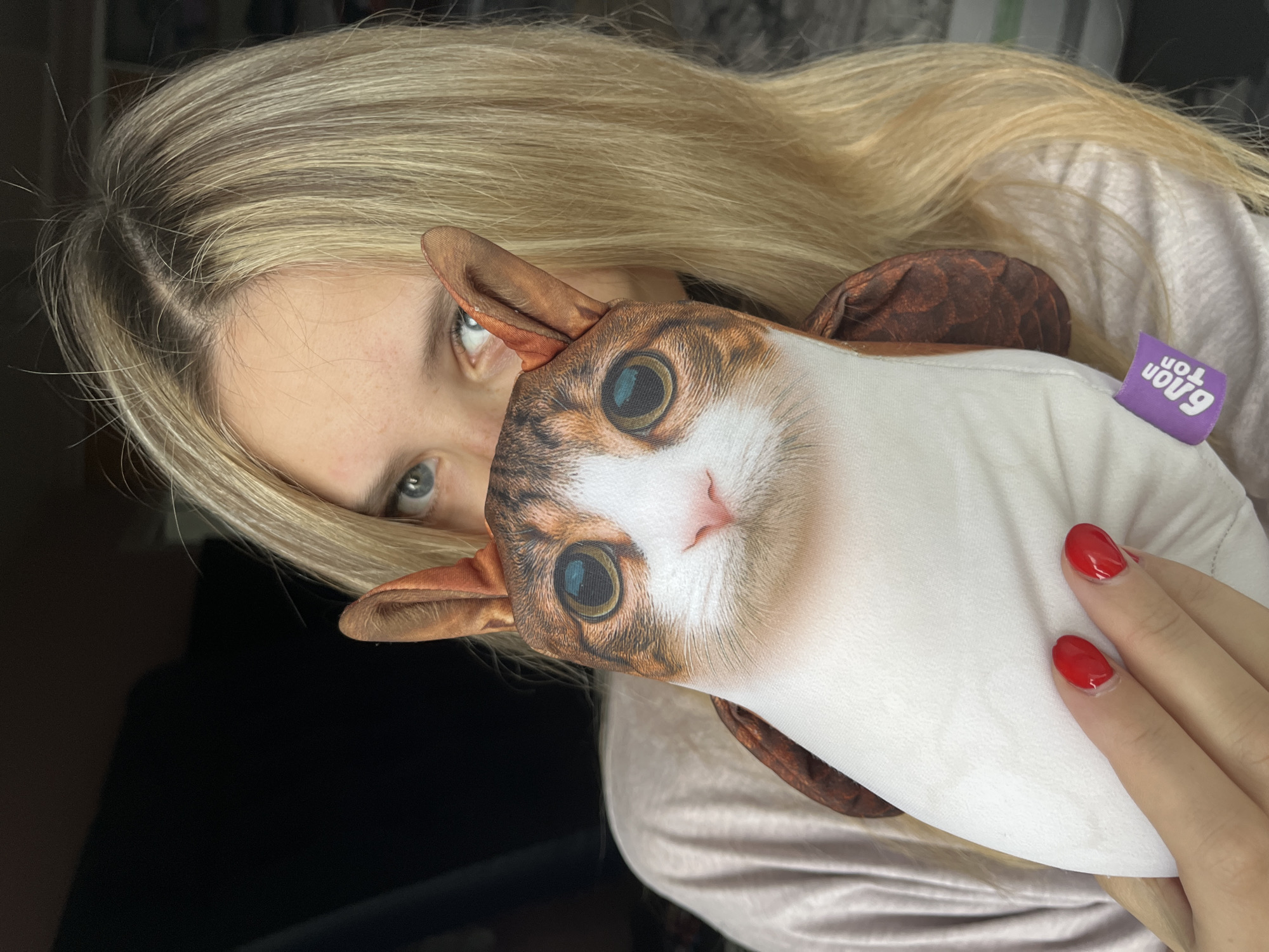 A young woman with blonde hair and blue eyes is holding a plush toy of a cat's face in front of her own. The toy is white with brown and orange markings on the cat's face, including large, detailed eyes and a pink nose. The woman is wearing a light-colored shirt, and her fingernails are painted red. A small purple tag with white lettering is visible on the plush toy. The background is dark, suggesting an indoor setting, possibly near a window. The image was taken in the city of Chaikovsky, Russia.