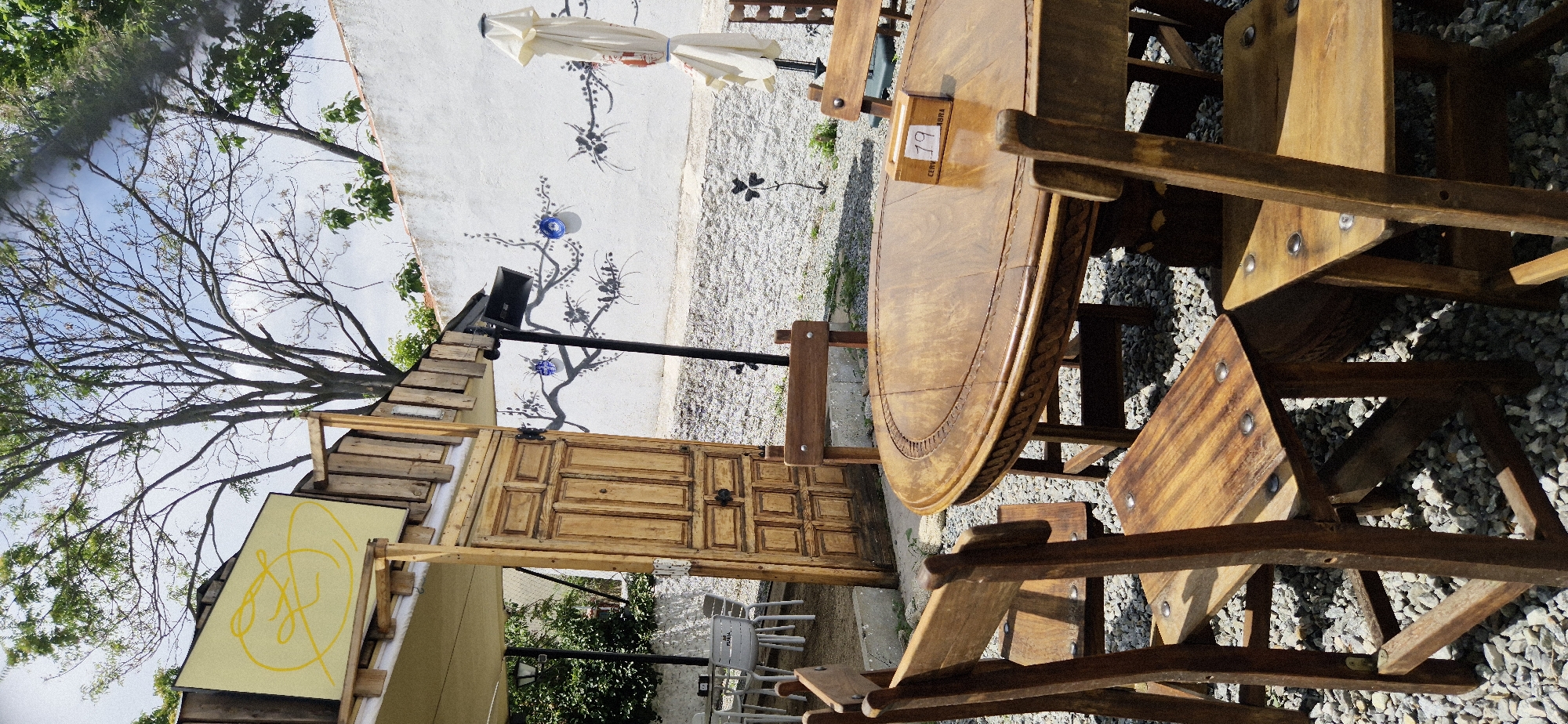 The image depicts an outdoor seating area with rustic wooden furniture, possibly in a cafe or restaurant setting. A round, intricately carved wooden table is the centerpiece, surrounded by several wooden chairs and benches. A small white sign with the number "19" is affixed to the table.
In the background, a white wall is adorned with a mural of a bare tree with dark branches and subtle floral accents. A blue decorative object, possibly a ceramic plate or bowl, is mounted on the wall near the mural. An umbrella is positioned behind the table, partially obscuring the wall.
To the left of the table, there's a wooden structure that appears to be a counter or serving area, constructed from rough-hewn planks. A large, old wooden door with multiple panels stands open within this structure. Above the door, a yellow board with a stylized yellow drawing is visible.
A bare tree with intricate branches dominates the left side of the frame, its silhouette contrasting with the bright sky. The ground is covered in small gray gravel. Several white plastic chairs are visible further in the background, suggesting additional seating.
The lighting indicates it is daytime, likely sunny, with strong shadows cast by the furniture. The overall impression is one of a casual, slightly weathered, and artistic outdoor space. There is no visible indication of people or a specific event taking place.