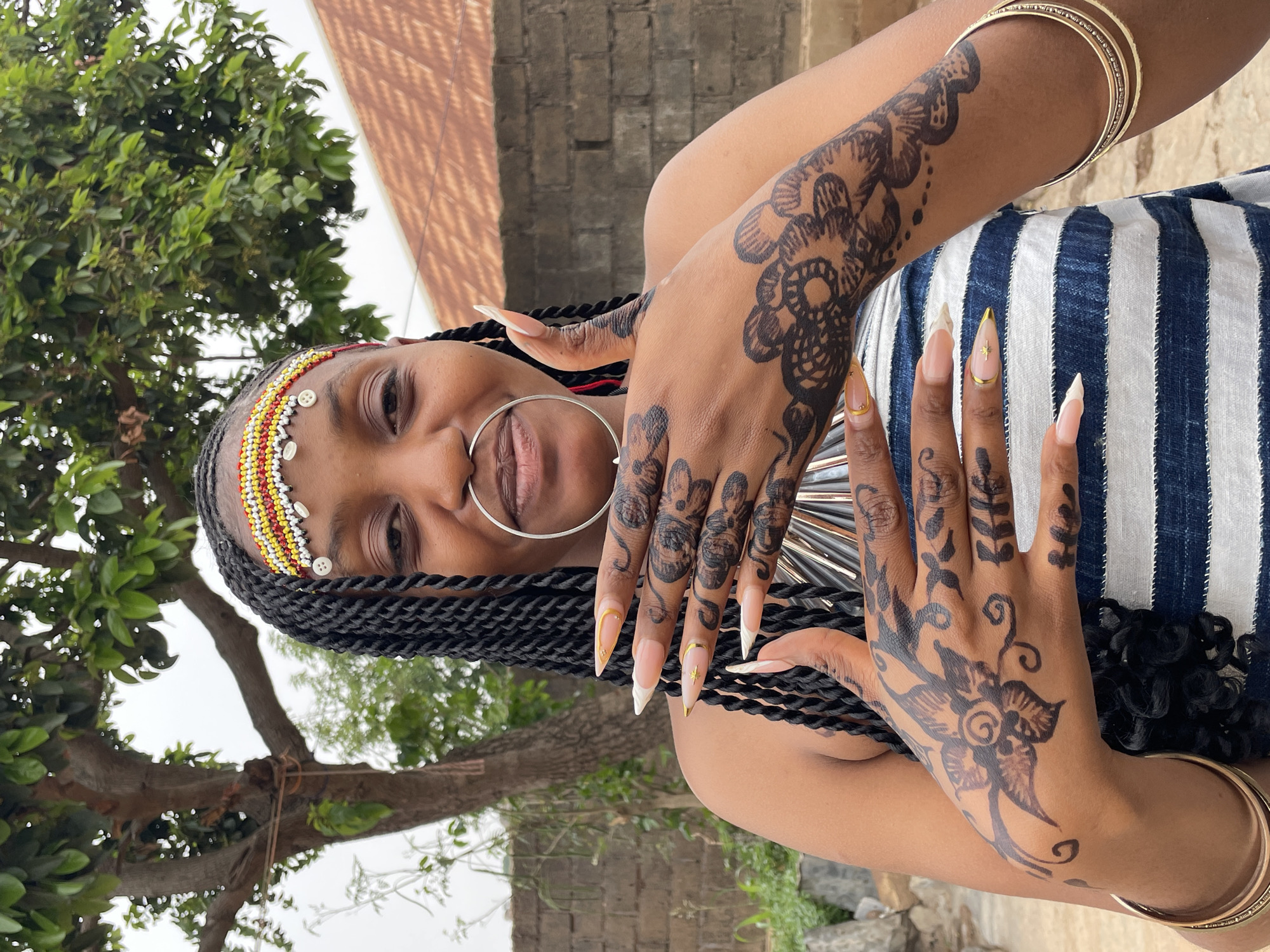A woman poses in front of a blurred background featuring a tree, wall, and roof, likely in Jos, Nigeria. She wears a beaded headband with white button-like ornaments, and a large silver nose ring. Her hands are adorned with intricate henna patterns. She is wearing long, decorative nails. Gold bangles are on her arms. She is wearing a striped blue and white garment.