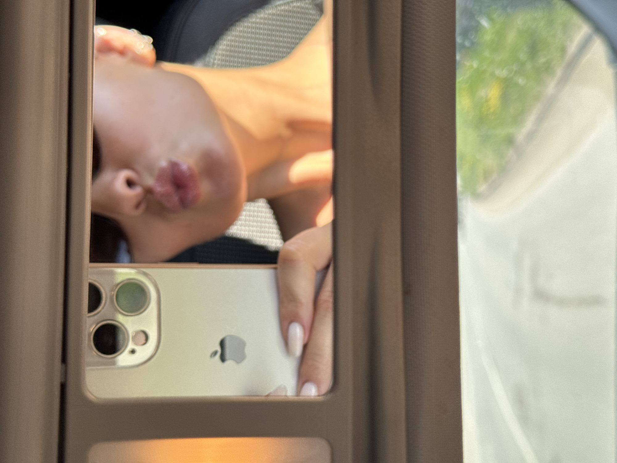 A selfie of a person is taken using a cell phone camera in a car sun visor mirror. The person is tilted to the side with pursed lips. The cell phone is silver and has three lenses. A hand is holding the cell phone. The surrounding environment includes the car interior and a blurry view through the car window with greenery outside. The location cannot be determined.