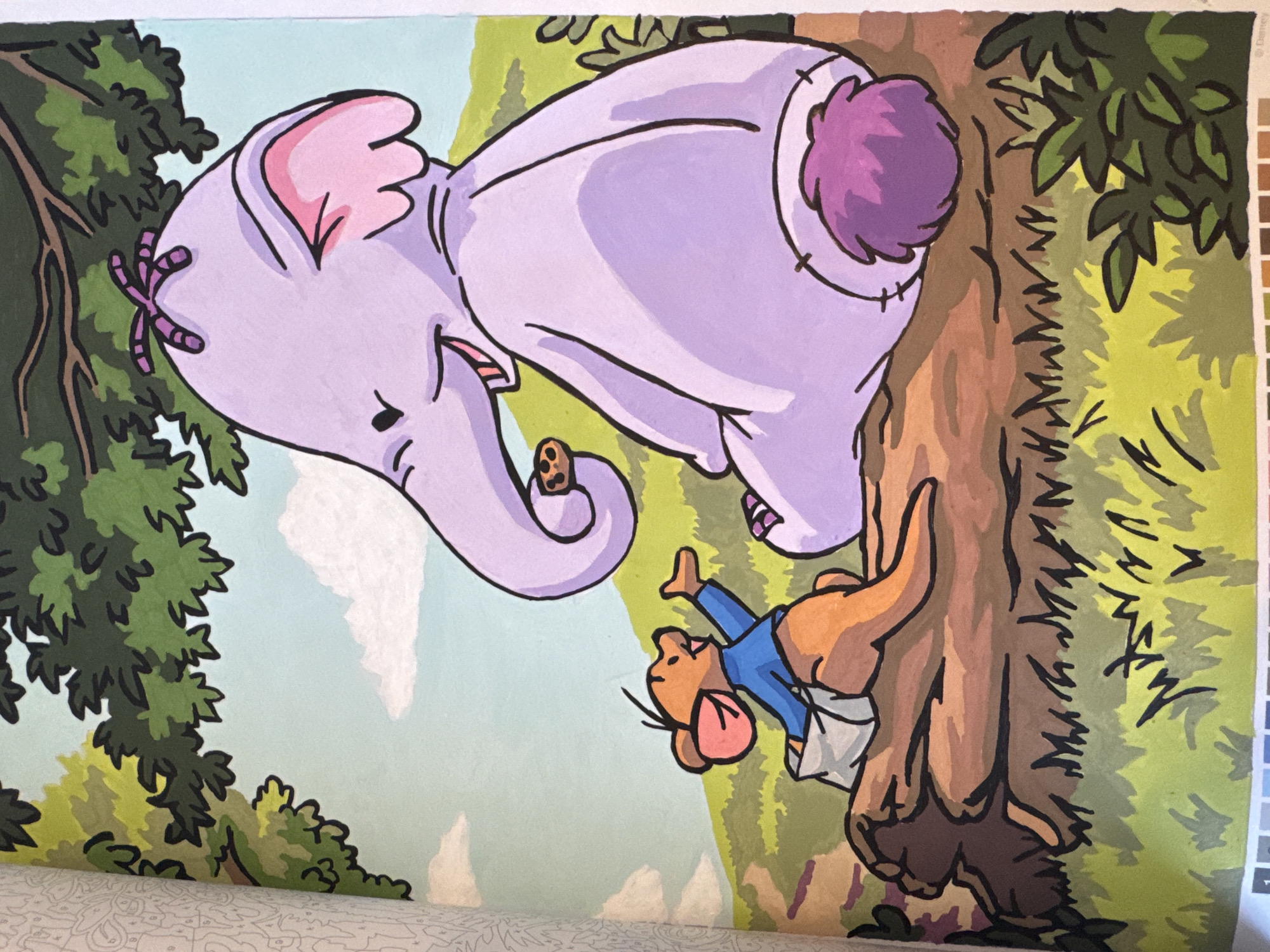 An outdoor scene depicts a purple elephant and a kangaroo sitting on a log. The elephant is larger, with large ears, a trunk holding a cookie, and a purple tuft of hair on its head. It is looking towards the kangaroo. The kangaroo is smaller, wearing a blue shirt and a diaper. It is reaching out towards the elephant. The background shows green trees, a light blue sky with white clouds, and green grass. The scene is illustrated in a cartoonish style.