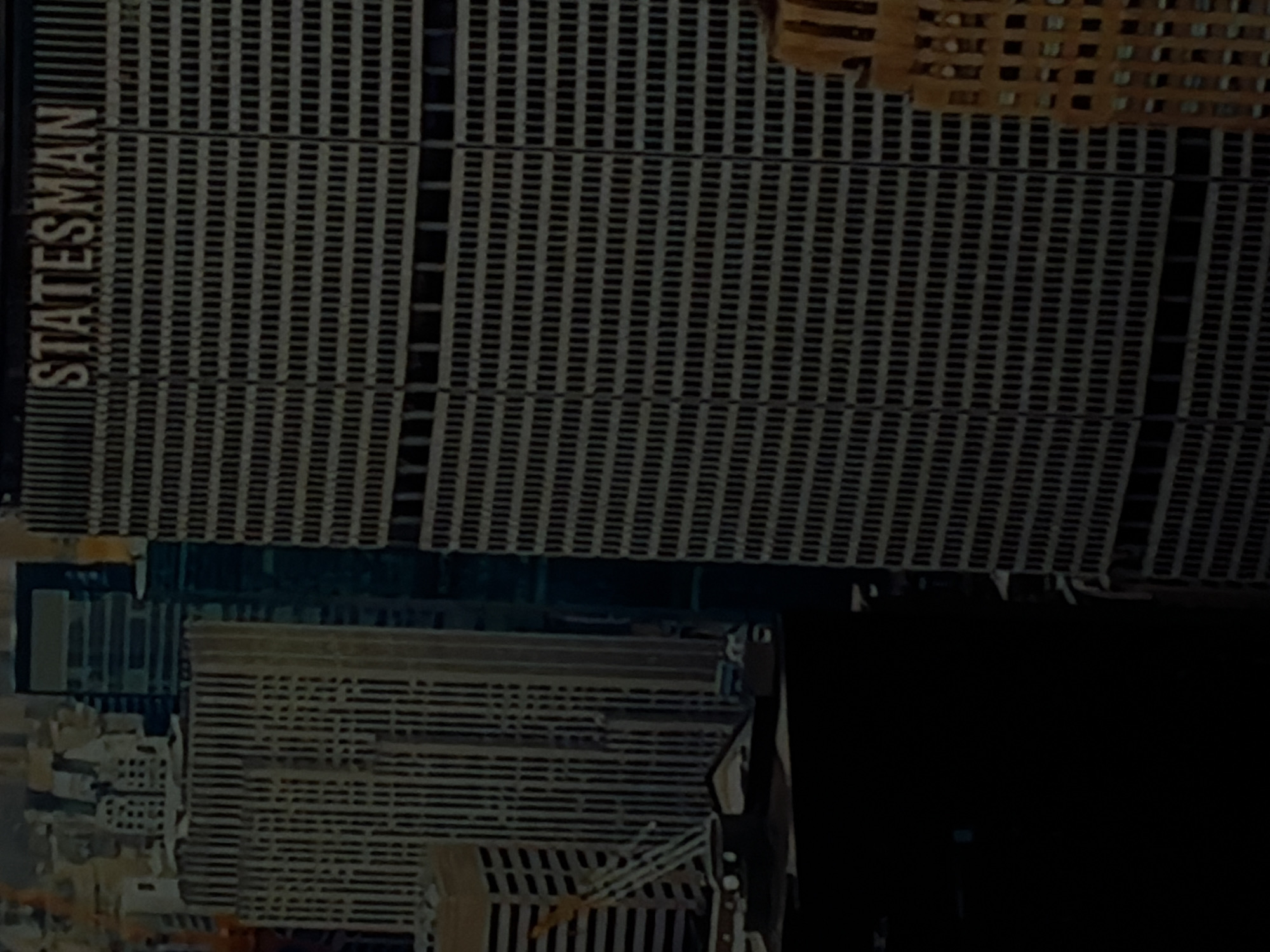 This rotated image captures a dark, urban scene, likely set in Sofiivska Borshchahivka, Ukraine. The primary subject is a large, imposing building with a distinctive, finely gridded facade composed of numerous repetitive vertical and horizontal lines in light gray against dark recesses. The word "STATESMAN" is clearly visible in tall, vertical block letters on the left side of this building.
The overall lighting is very dim, suggesting either nighttime or extremely low-light conditions, with the scene appearing largely desaturated and dark. To the right, another building or section of a building with a similar gridded pattern but in a warmer, brownish tone is partially visible. Further down, other structures are faintly discernible amidst deep shadows, contributing to a dense city landscape. No people or specific activities are visible. The atmosphere conveyed is stark and architectural, dominated by the geometric patterns of the buildings under subdued lighting.