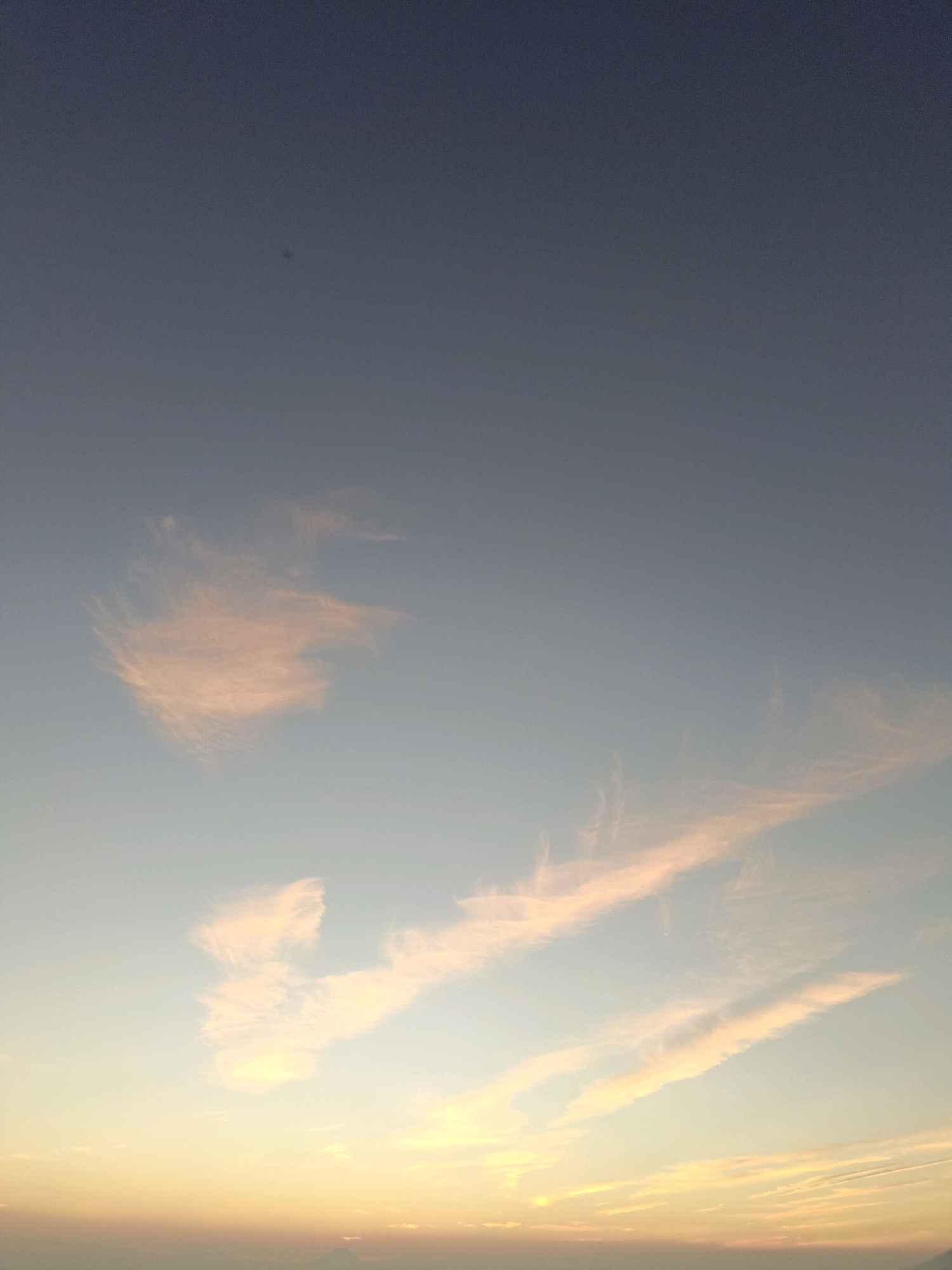 The image shows a sky with wispy clouds tinged with orange and pink, indicative of sunrise or sunset. The sky transitions from a deep blue at the top to a lighter blue and then yellow towards the horizon. In the lower portion of the image, there appears to be a faint, dark silhouette of land or buildings against the bright horizon. There are no people or specific identifiable objects in the image other than the clouds and the sky. The location is specified as Butuh Kidul, Indonesia.