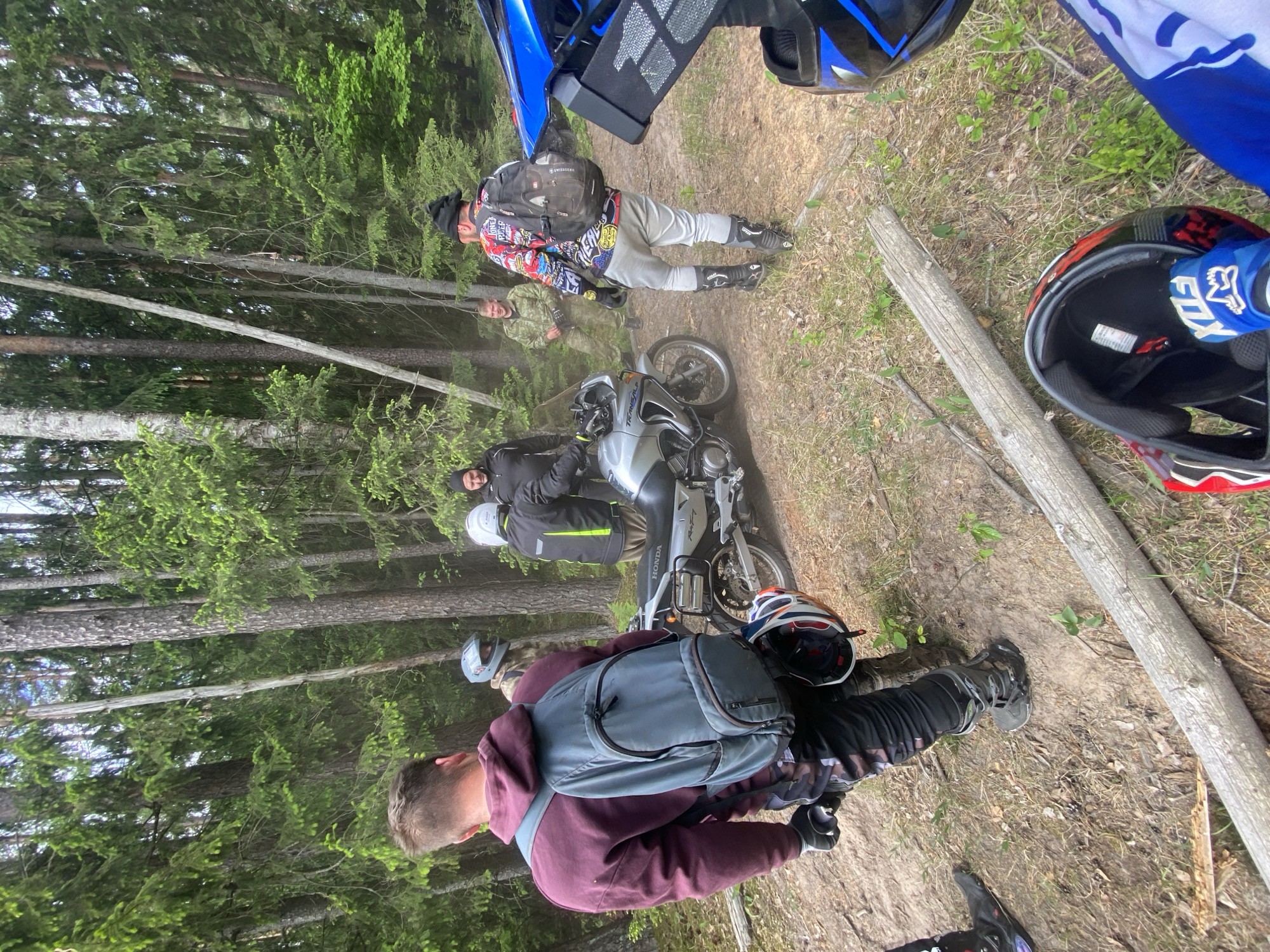 A group of people are gathered on a dirt path in a wooded area. The path is lined with tall trees. A motorcycle is parked on the path. Several people are standing near the motorcycle, while others are further down the path. A log lays near two motorcycle helmets, which are near a patch of green vegetation. The scene is located in the Парэцкі сельскі Савет, Belarus.