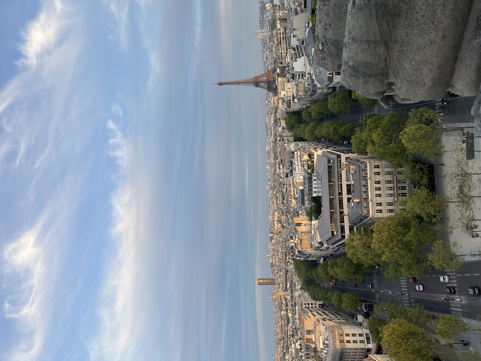This media file presents a panoramic, high-angle view of the city of Paris, France, under a clear, partly cloudy sky. The perspective suggests the photo was taken from an elevated structure, possibly a monument or a tall building.
In the upper third of the image, the sky transitions from a lighter blue near the horizon to a deeper blue overhead, adorned with scattered white, wispy cirrus clouds. The lighting suggests late afternoon or early evening, with soft sunlight illuminating parts of the cityscape.
The iconic Eiffel Tower stands prominently in the mid-ground on the left, a dark, elegant silhouette against the brighter sky. To its right and extending into the distance, the dense urban landscape of Paris unfolds, characterized by numerous multi-story buildings with classic Haussmannian architecture, featuring light-colored facades and grey roofs.
A wide, tree-lined boulevard, likely one of Paris's grand avenues, cuts through the city in the lower portion of the frame. Lush green trees form a continuous canopy along its sides. Several cars are visible on the road, indicating active traffic; some vehicles appear white, black, or red, and one resembles an emergency vehicle.
In the far distance on the right, the distinct, tall, rectangular form of the Montparnasse Tower can be seen, its upper section appearing to catch the last rays of sunlight with a golden hue. On the immediate right side of the foreground, a large, textured grey stone architectural detail, possibly part of a balustrade or a decorative element of the vantage point, partially frames the scene.
No discernible text is visible within the image. The overall impression is one of a vast, bustling European capital bathed in the tranquil light of approaching dusk.