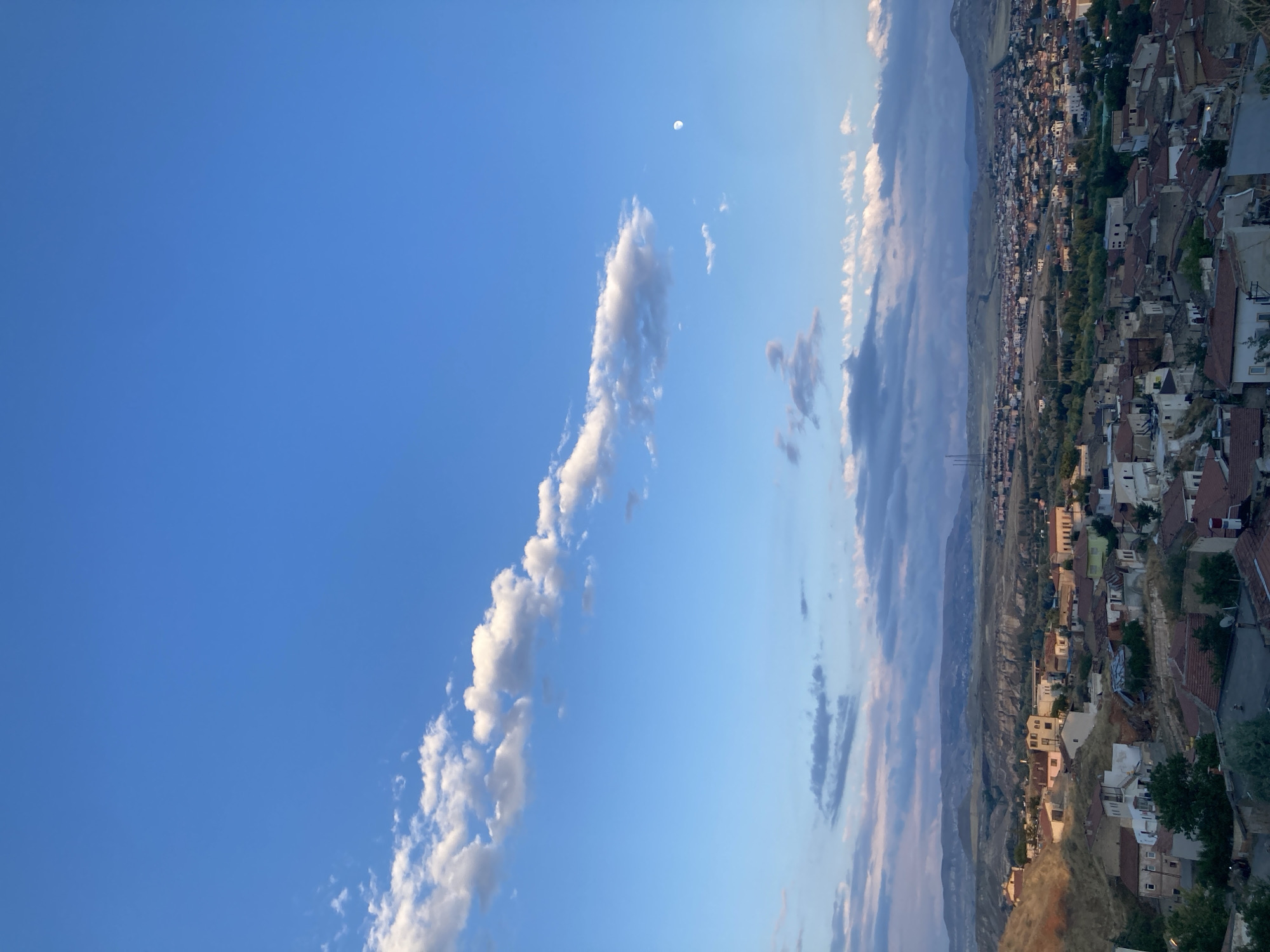 The image captures a panoramic view of Avanos İlçe Merkezi, Turkey, likely during the late afternoon or early evening, as indicated by the soft, warm light bathing the landscape and the visible crescent moon in the sky. The foreground is dominated by a densely packed collection of buildings with terracotta tile roofs, characteristic of Mediterranean architecture. These structures are nestled into the hilly terrain, with some appearing to be older, more weathered buildings on a steeper incline, and others, possibly newer constructions, spread across the flatter areas.
The midground reveals a more extensive urban sprawl, with a continuous tapestry of houses and buildings extending towards the horizon. Densely planted trees are interspersed throughout the town, creating pockets of greenery. In the background, rolling hills and mountainous terrain form a dramatic backdrop. The geological formations suggest a dry, arid climate, with exposed rock faces and sparse vegetation on the slopes. The sky is a vibrant blue, dotted with wispy white clouds that are beginning to catch the warm hues of the setting sun. There are no visible people or animals in the frame. The overall impression is one of a well-established town situated within a rugged, natural landscape.