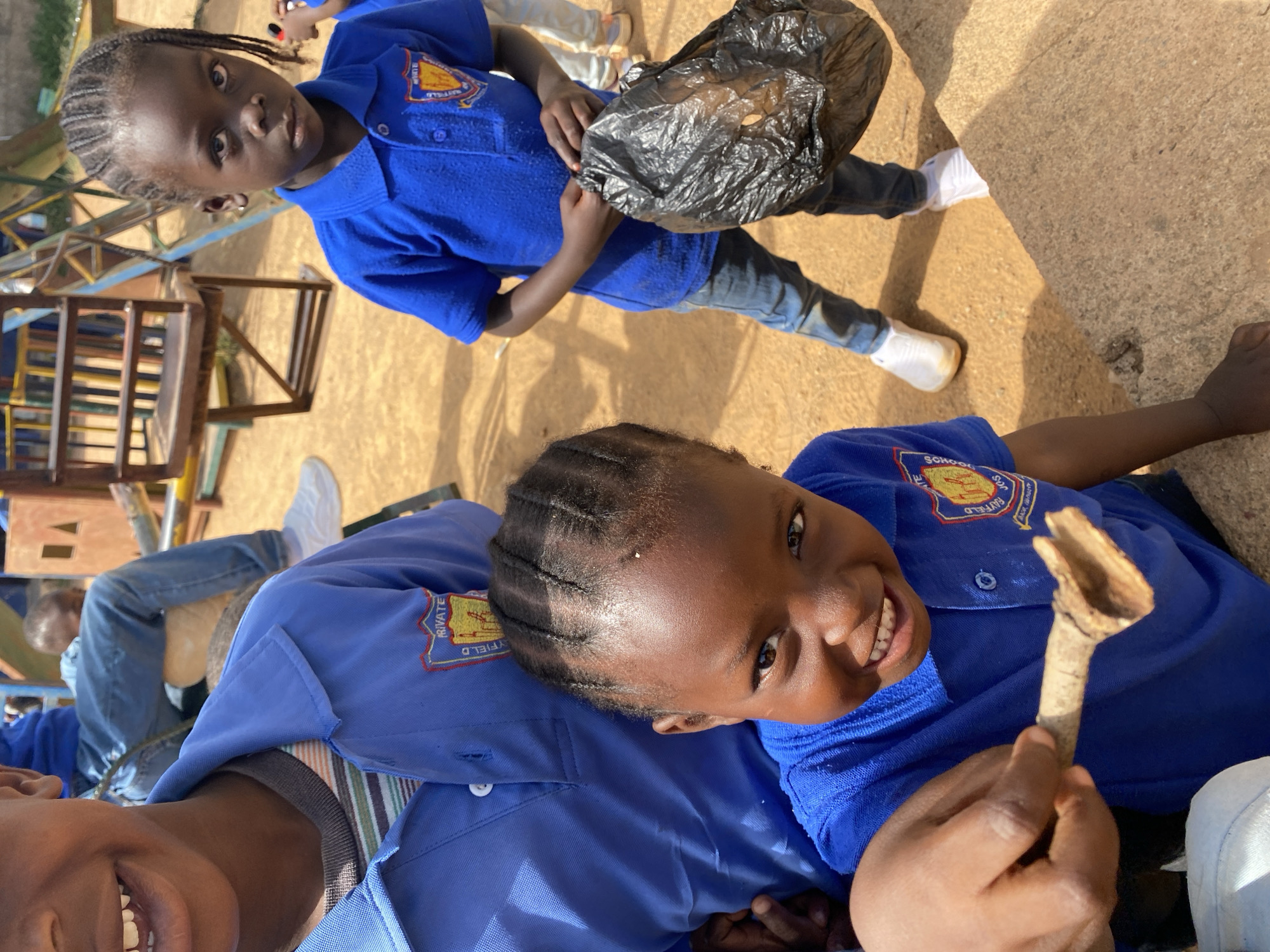 In Gura Top, Nigeria, a group of children wearing blue school uniforms are outdoors. One child holds a black trash bag, wearing white sneakers and jeans. Another child holds a small wooden stick. A building structure is visible in the background. The environment appears to be a yard with a dirt surface.