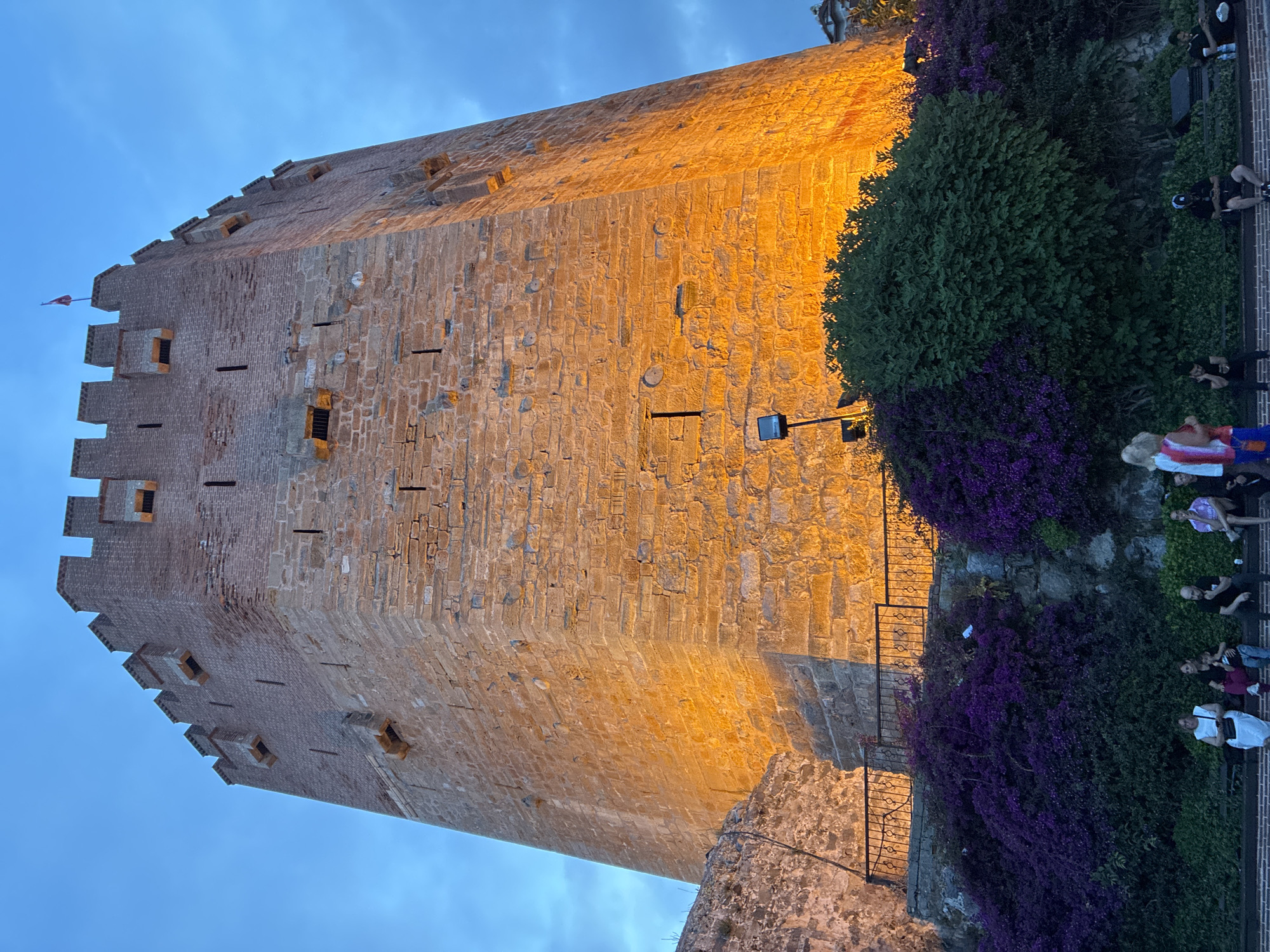 This image captures a majestic, ancient stone tower, likely the Red Tower (Kızıl Kule) in Alanya, Turkey, dramatically illuminated against a twilight sky. The tall, broad stone tower, constructed from rough-hewn stone blocks, stands prominently, showcasing its battlements along the top edge. Small, rectangular window openings and arrow slits are visible across its sturdy walls, some containing dark bars. A flag, possibly Turkish, flutters from a mast near the tower's summit.
The tower's surface glows with a warm, golden light, emanating from strategically placed floodlights visible at its base, creating a striking contrast with the deep blue and subtly cloudy twilight sky above. At the base of the tower, on the right side, a vibrant cascade of purple bougainvillea-like flowers and lush green foliage grows along a stone wall. A dark metal railing and some stone steps are also present. Several people are scattered in this area, appearing as visitors or tourists. Some are standing, others are seated on low walls or benches, observing the illuminated landmark or enjoying the evening atmosphere. The scene conveys a serene and appreciative ambiance, highlighting the historical grandeur of the site at dusk. No discernible text is visible within the frame.