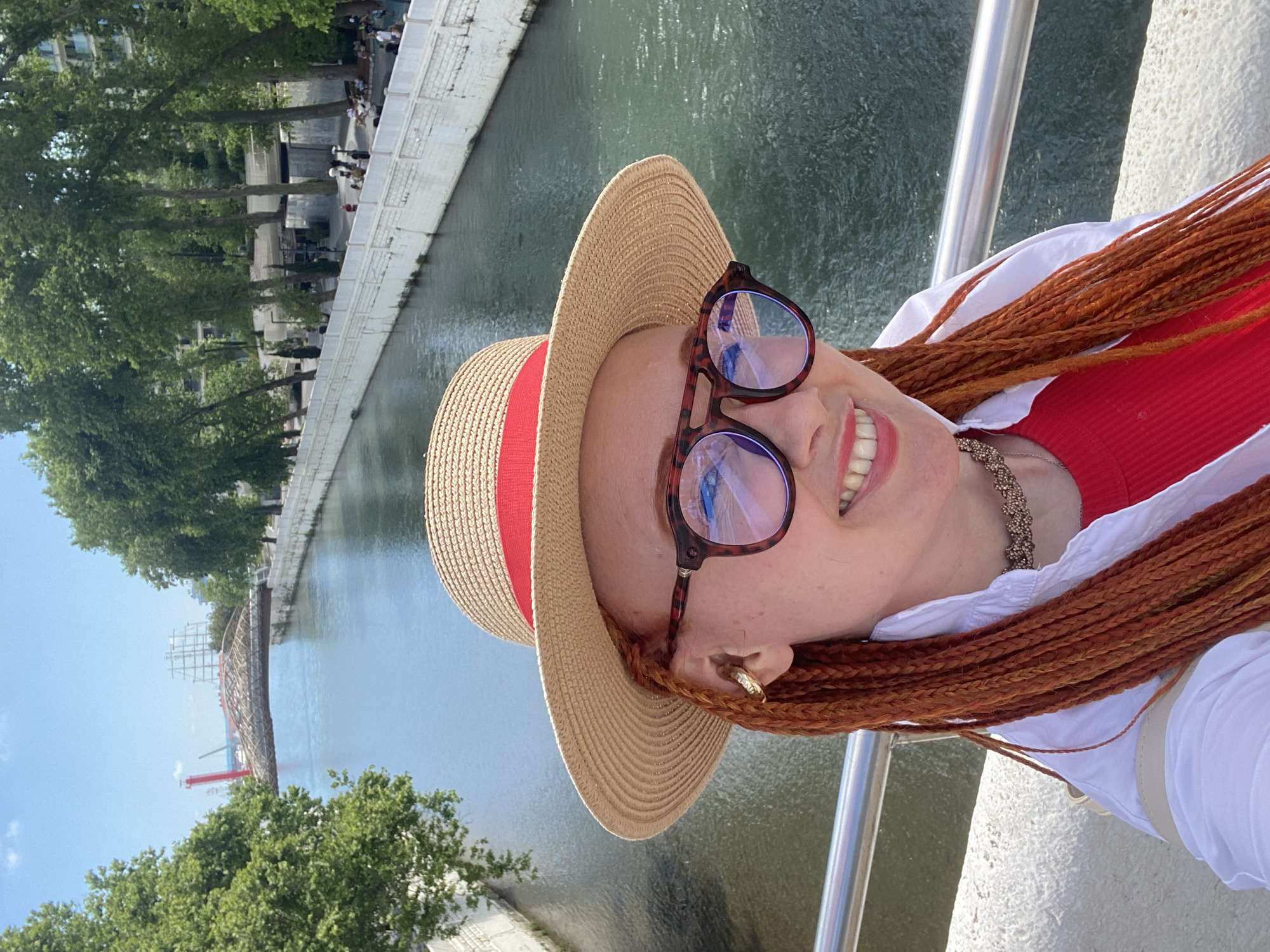 A woman with red braided hair, wearing a straw hat with a red ribbon, glasses, and a red top with a white shirt, takes a selfie on a bridge over a canal in Sochi, Russia. In the background, a distinctive bridge with a curved wooden structure spans the canal, with trees and buildings lining the banks. The sky is blue with some clouds.