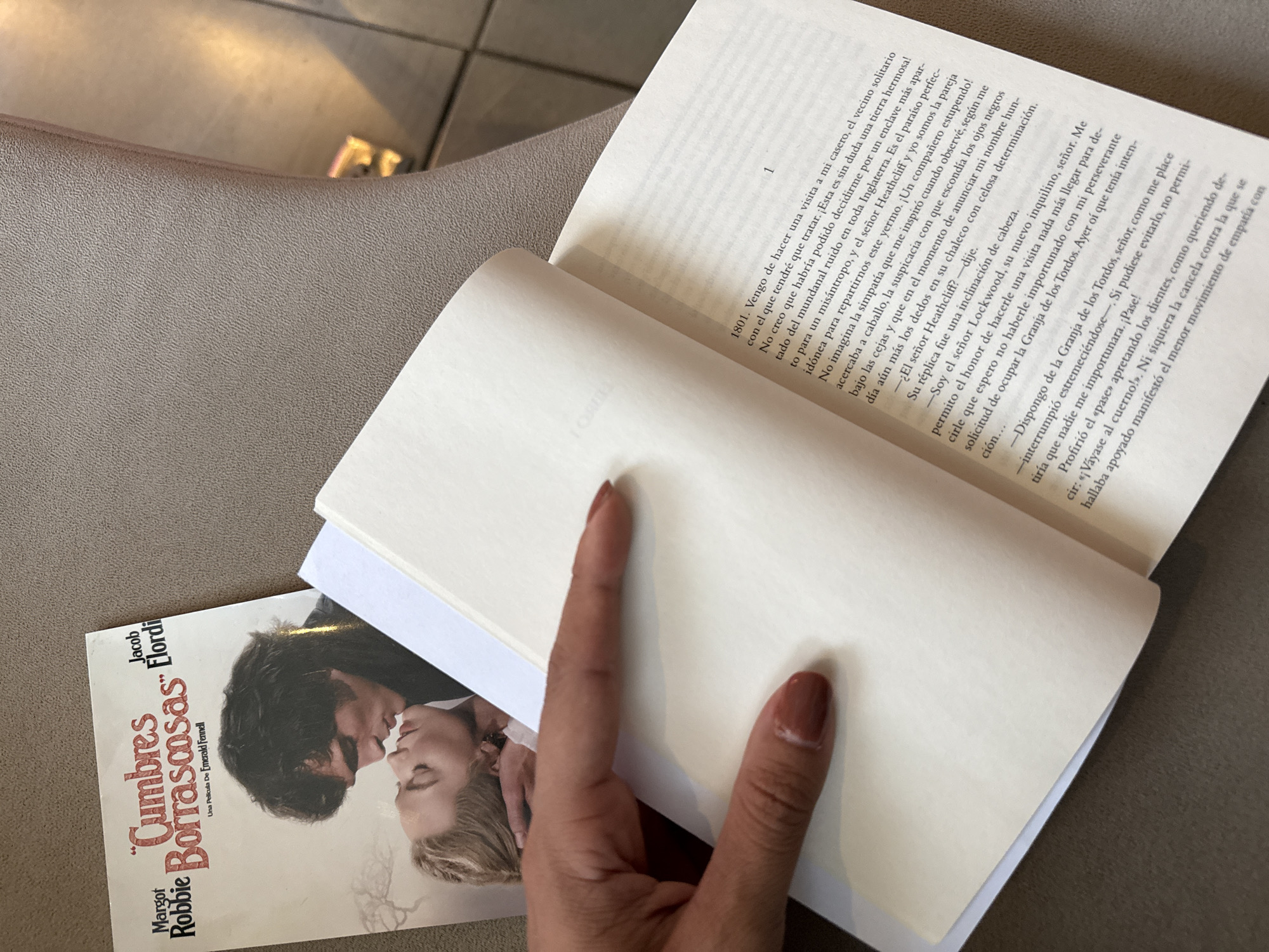 Here is a detailed description of the image:
**Scene Description:**
The image shows an open book resting on a surface, with a hand holding open the pages. The book on top appears to be a Spanish language copy of "Wuthering Heights," based on the visible cover. The book's open pages display text in Spanish. Below this book, there's a Spanish version of Wuthering Heights by Emily Brontë, with actors Margot Robbie and Jacob Elordi on the cover.
**Objects:**
* Book (Spanish edition of "Wuthering Heights")
* Open Book
* Hand
**Actions:**
* Holding a book open.
* Reading (implied).
**Environment:**
* The surface appears to be a neutral, possibly indoor setting, potentially inside a building.