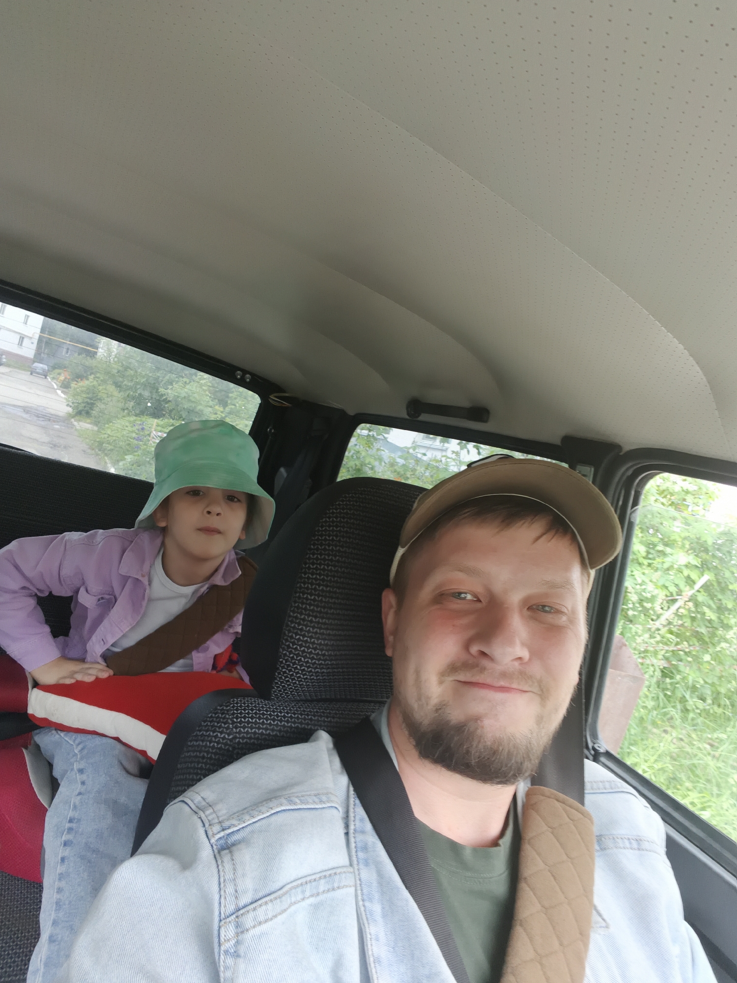This image depicts a selfie taken inside a vehicle in the city of Chaikovsky, Russia. The foreground features an adult male, presumably the driver, with light skin, a brown beard, and short hair, wearing a light brown baseball cap, a green t-shirt, and a light blue denim jacket. He is secured by a black seatbelt, which includes a quilted brown shoulder pad, and is smiling faintly while looking directly at the camera. In the background, a child is seated in a rear passenger seat, occupying a red and white booster cushion. The child is wearing a light green bucket hat, a light purple long-sleeved shirt, and light blue jeans, with a brown strap across the chest. The child maintains a neutral expression and looks toward the camera. The vehicle's interior features a light gray, perforated headliner and dark, textured fabric seats. Through the rear window, a wet paved road, a multi-story light-colored residential building, and green trees and bushes are visible. Another vehicle can be seen in the distance on the road. The side window on the right reveals dense green foliage, suggesting an outdoor environment with significant vegetation under an overcast sky.