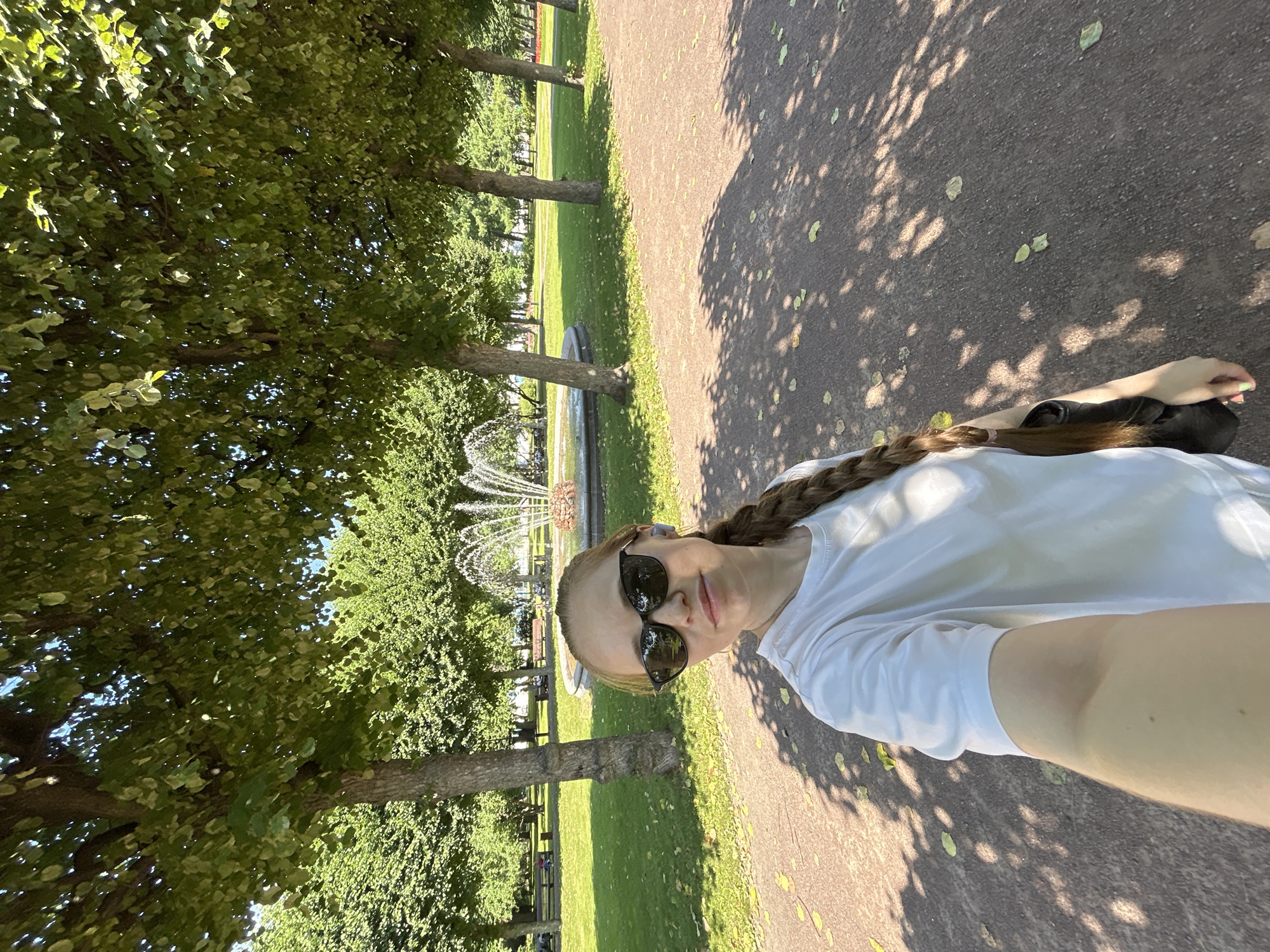 A young woman with long braided brown hair and sunglasses takes a selfie in a park on a sunny day. She is wearing a white t-shirt and a black bag is visible over her shoulder. In the background, there is a fountain, green grass, trees, and park benches. The scene appears to be taken in the Summer Garden in Saint Petersburg, Russia, identifiable by the architectural style of the fountain and the manicured park environment characteristic of the city. The sunlight casts dappled shadows from the trees onto the pathway.