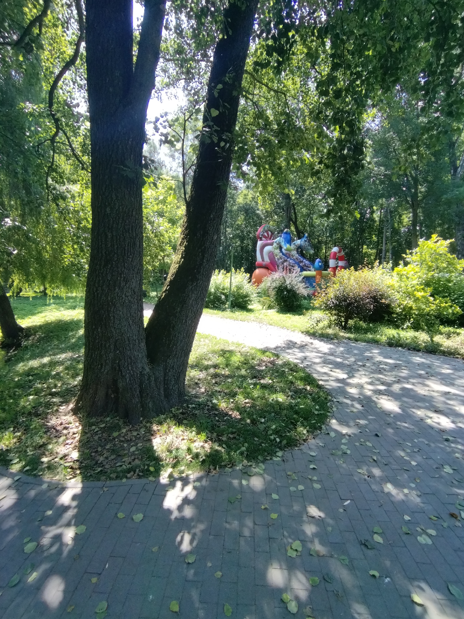 This image depicts a park scene in Khmelnytskyi, Ukraine, on a sunny day. In the foreground, a paved walkway with scattered leaves runs through the park, bordered by grass and trees. The path is dappled with sunlight and shadow cast by the overhead foliage. To the left, the thick trunks of two large trees dominate the view, with lush green leaves and branches creating a canopy above. In the background, an inflatable playground structure, brightly colored with blue, red, and white elements, is visible amidst greenery. The overall environment suggests a recreational area within a city park.