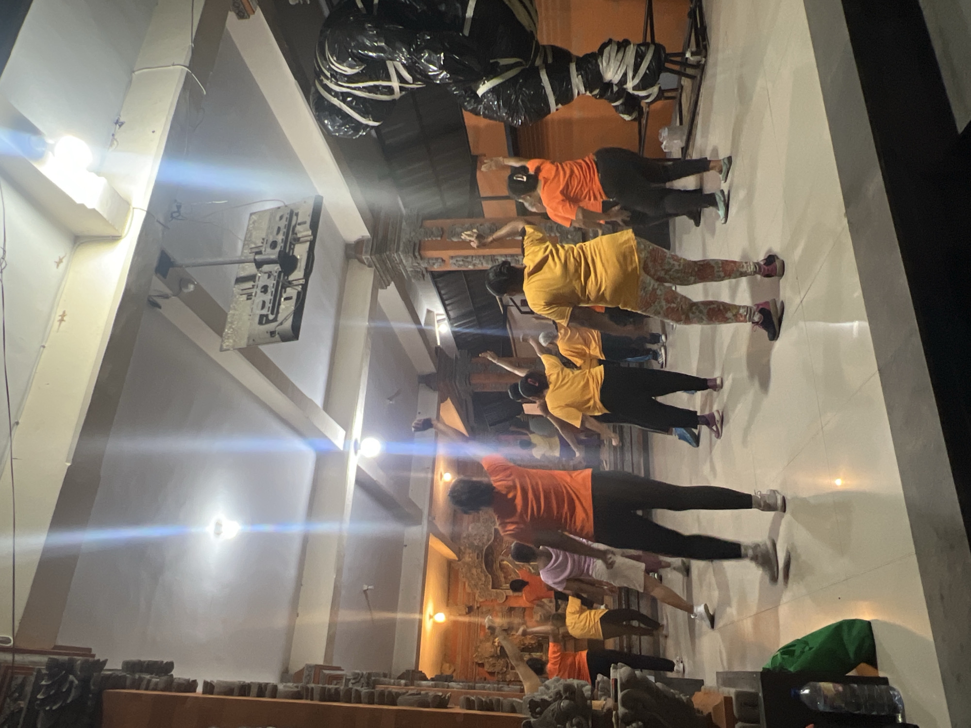 A group of people are participating in a yoga class indoors in Ubud District, Indonesia. The individuals are dressed in a variety of colors, including orange, yellow, and black, and are standing in rows on a tile floor. They are all performing a similar pose with their arms extended. The room has high ceilings with exposed beams and overhead lighting. There is a structure wrapped in black and white material suspended from the ceiling, and a display of carved wooden panels can be seen in the background.