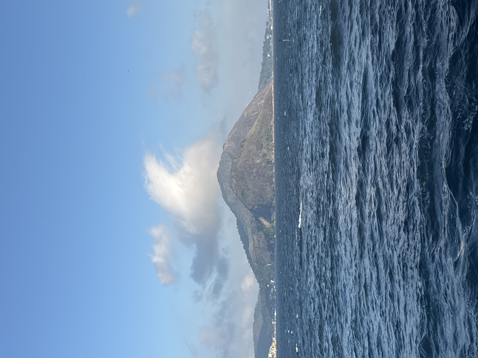 The image depicts a wide shot of the ocean in front of a mountainous landscape. The water is a deep blue with whitecaps, indicating moderate waves. In the background, a large, rounded mountain dominates the scene, with green vegetation covering its lower slopes. A few clouds are visible in the clear blue sky. On the left side of the image, a coastline with buildings can be seen, suggesting a city or town. On the right side, a white buoy stands in the water. This scene is characteristic of the view of the Sugarloaf Mountain (Pão de Açúcar) from Niterói, Brazil, across Guanabara Bay. There are no people or explicit actions visible in the image.
