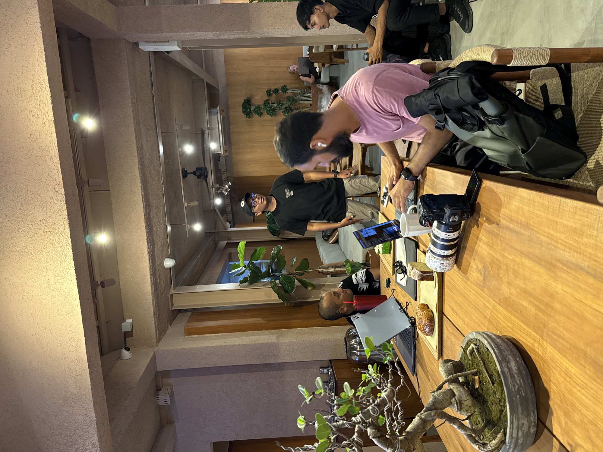 The image captures an indoor scene, likely a cafe, co-working space, or casual meeting area, characterized by warm wooden tables and structural elements, complemented by textured beige walls and ceiling. Multiple individuals are present, engaged in various activities.
In the foreground, a man with a beard and wireless earbuds, wearing a pink t-shirt and a smartwatch, leans over a wooden table, actively interacting with a device. Next to him on the table are a black camera with a large lens, a black backpack, and a white mug. To the left, another man in a black t-shirt, khaki pants, glasses, and a black cap stands, looking ahead. Further back, a third man in a black t-shirt is partially visible. In the mid-ground, a man in a red shirt is seated, using a laptop and holding a red cup with a straw. Another man in a black t-shirt is seen in the upper background.
The table also holds other items, including an open laptop, a mobile phone, black-rimmed glasses, a pastry or bread item on a small tray, and a slice of cake. A small bonsai tree in a grey pot adds a decorative touch. In the background, potted plants contribute to the ambiance. The area is illuminated by several ceiling spotlights, some creating glare. Security cameras are mounted on the walls.
The scene suggests a collaborative or individual work session, possibly involving photography or content creation, given the visible camera and multiple laptops. The presence of food and drinks indicates a relaxed environment where people are working or socializing. The context provided specifies the location as North Kuta, Indonesia. No specific legible text is visible beyond potential brand names on devices, like "CANON" on the camera lens, but the text is not clearly readable. The time of day is not discernible from the indoor lighting.