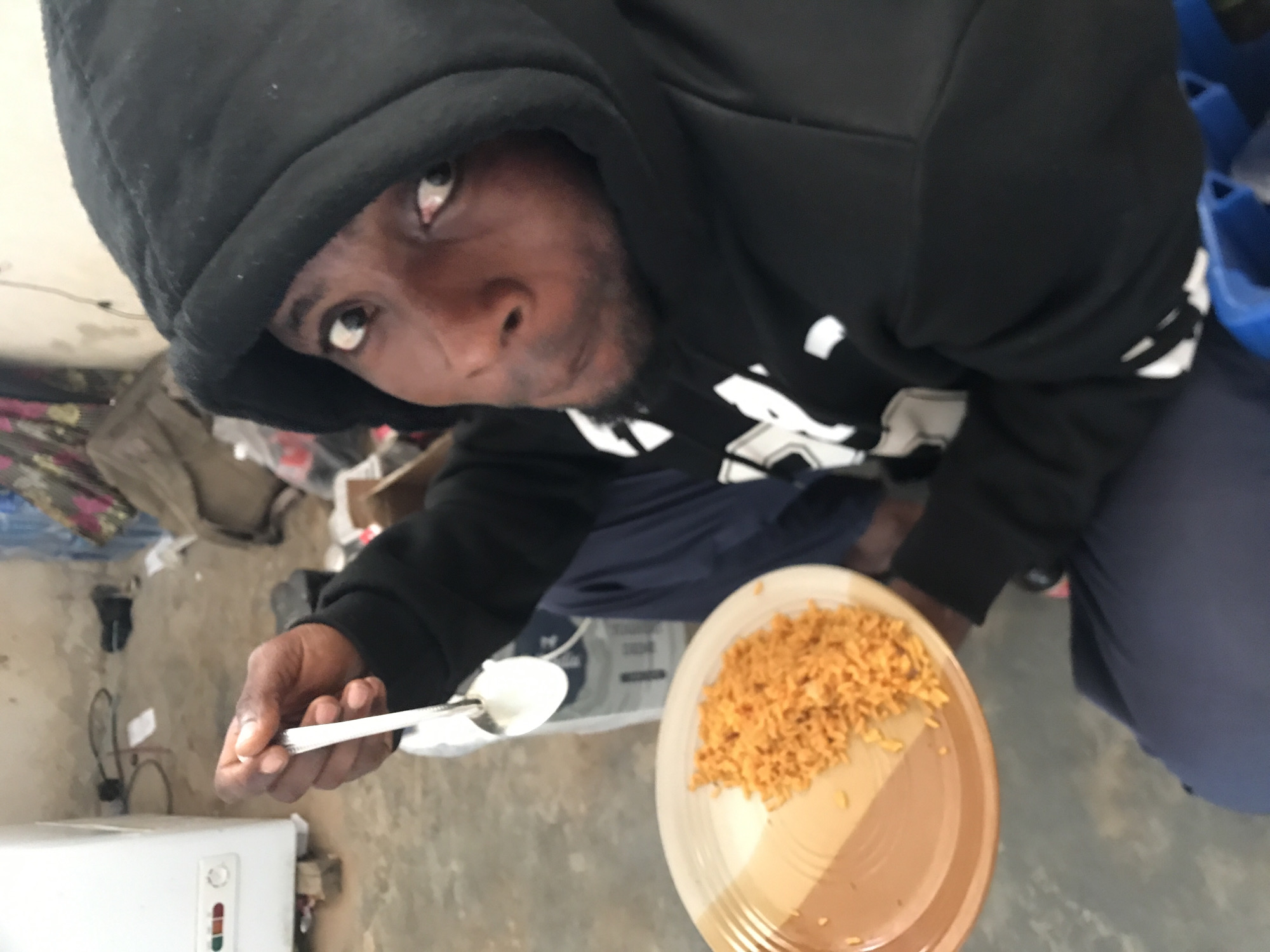 A dark-skinned man is depicted indoors in New Stadium, Nigeria, looking directly at the camera with a wide-eyed gaze and a slight pout. He is wearing a black hooded sweatshirt with the hood up and dark blue pants. In his raised right hand, he holds a silver-colored spoon containing a small amount of white, pasty substance. His left hand supports a round, two-toned plate (brown and white) filled with a pile of orange-colored rice. The background features a concrete or tiled floor. To the man's left, a white upright appliance, possibly a freezer or refrigerator, is visible, elevated on wooden blocks. The overall environment appears functional and somewhat cluttered, with cables and various dark objects scattered on the floor and in the distant background.