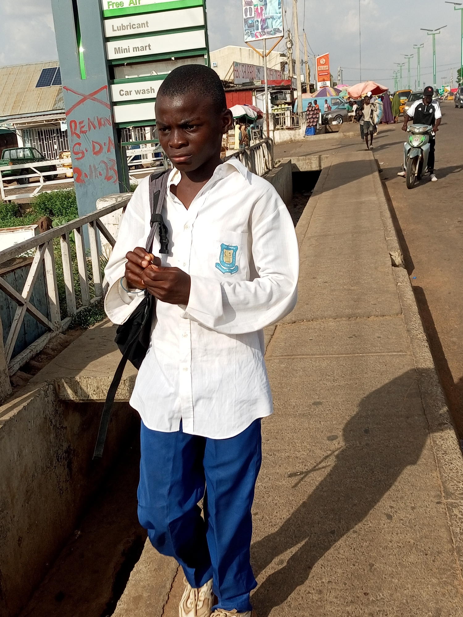 The image shows a young boy, likely a student due to his uniform, walking on a sidewalk in an urban environment. He is wearing a white long-sleeved shirt with a crest on the left breast and blue trousers. A backpack is slung over his right shoulder. The boy is looking down and to his left with a serious expression. The sidewalk is concrete and appears worn. To the left of the boy, there is a railing and a concrete structure that looks like a drainage ditch. In the background, there are buildings, shops, and a busy street with vehicles and other pedestrians. One of the shops has signage indicating "Free Air," "Lubricant," "Mini mart," and "Car wash." The overall scene suggests a bustling commercial area. The lighting indicates it is daytime, with shadows cast by the boy and other objects.