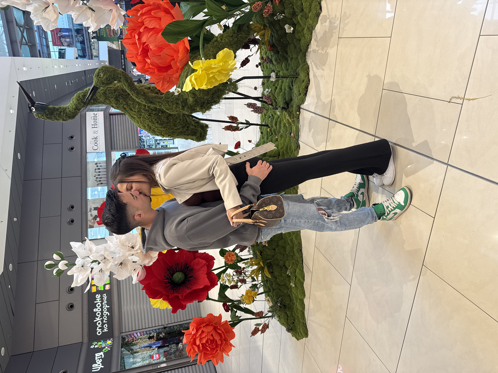 A couple is kissing in a mall in Sofia, Bulgaria. They are standing in front of a floral display with white, red, and orange flowers, and a large green bird sculpture. The woman is wearing a beige jacket and black pants, while the man is wearing a gray hoodie and ripped jeans. He has a Louis Vuitton purse.