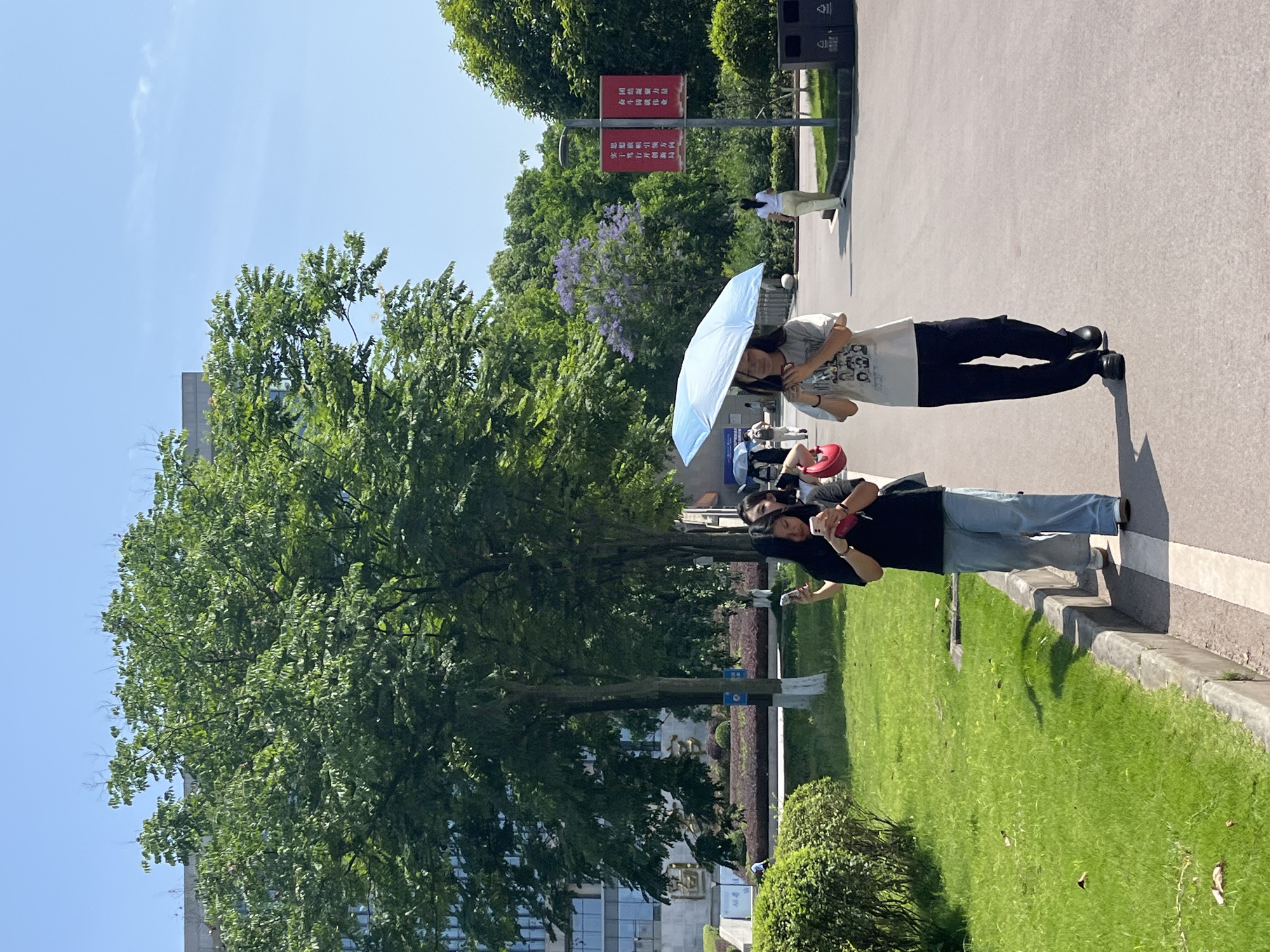 The image depicts an outdoor scene in Jiangjin, China. Two women are standing on a paved pathway beside a green grassy area. The first woman wears a black shirt and light blue jeans and is holding a phone in her hands. Next to her is a woman wearing a t-shirt and black pants, holding a light blue umbrella. Both appear to be looking at their phones. Large leafy trees provide a backdrop, with a building partially visible behind them. In the background, there is signage with Chinese characters and trash bins. The sky is clear and blue, suggesting it is daytime.