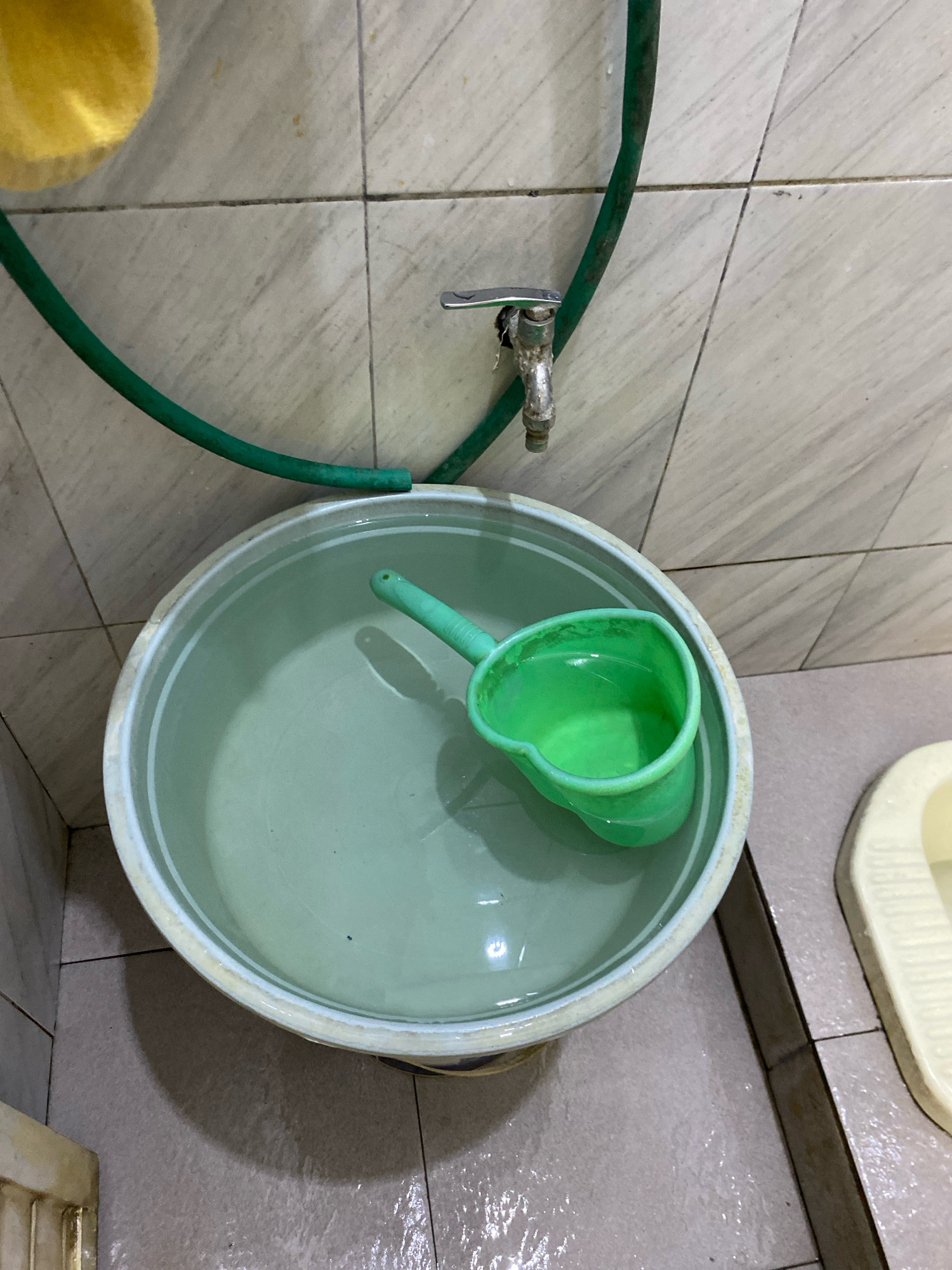 The image shows a bathroom scene in Gisik Cemandi, Indonesia. A large, light-colored basin filled with water is placed on the tiled floor. A bright green plastic ladle with water in it is resting inside the basin. A green hose is connected to a silver faucet mounted on the tiled wall above the basin. The floor tiles are light brown and appear wet in places. A portion of a light-colored toilet is visible to the right of the basin. A yellow object, possibly a sponge or cloth, hangs from the wall on the left.