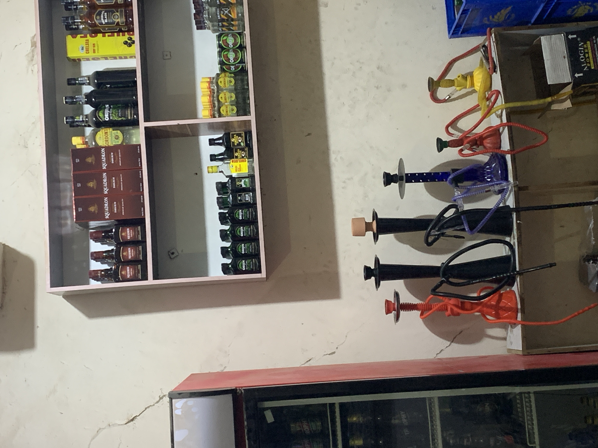 The image shows a liquor shelf stocked with various bottles of alcohol, including brands such as Chelsea Dry Gin, Gordon's, and Squadron, mounted on a white wall. Adjacent to the shelf are several disassembled hookahs with hoses and mouthpieces of different colors. Below, the top of a refrigerator or cooler unit is visible, its glass doors showing more beverages inside. The overall setting appears to be in a store or bar in New Stadium, Nigeria.