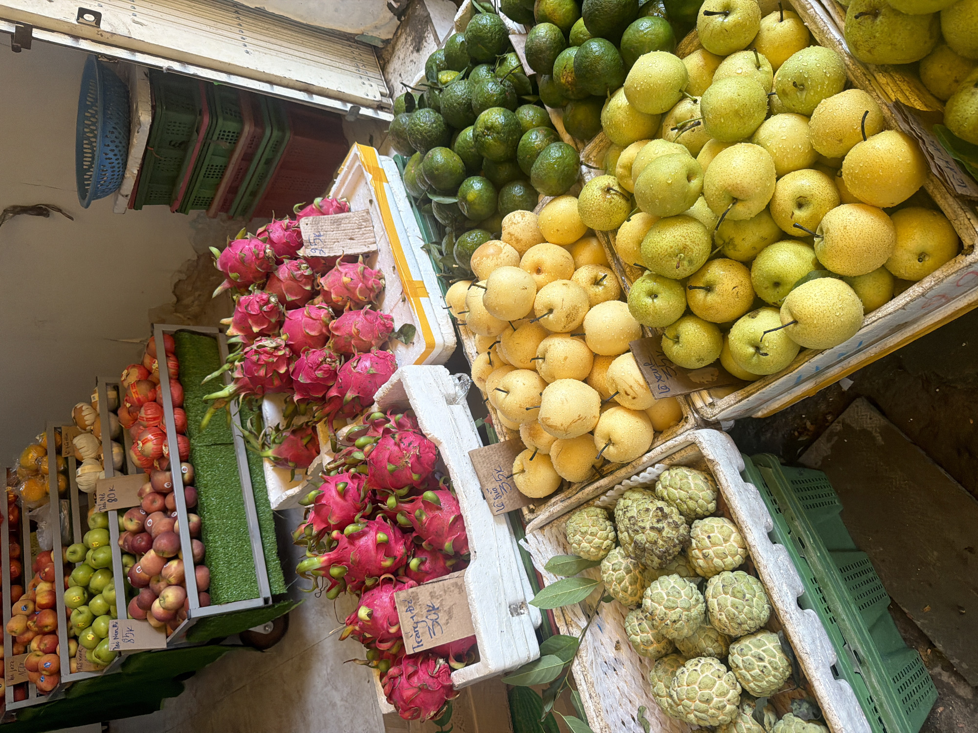 The image shows a fruit stand in Nha Trang, Vietnam, likely on a sidewalk or in a market. The stand features a tiered display of various fruits, including apples, dragon fruit, custard apples, pears, and possibly oranges or limes. Some of the fruit containers have handwritten price tags. In the background, stacks of plastic crates and a blue basket are visible, suggesting a commercial setting. The lighting suggests it is daytime. There are no people visible in the image.