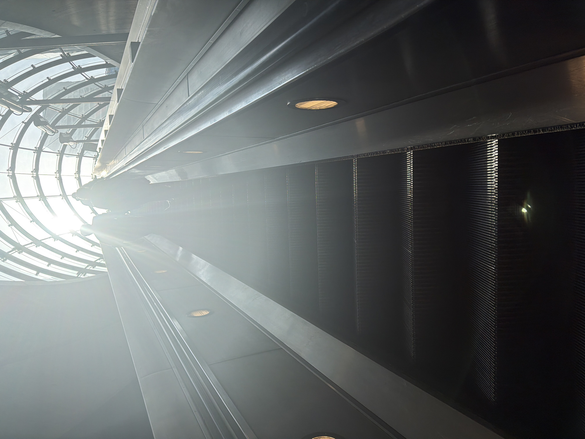 The image depicts the interior of an escalator at the Leadenhall Building (also known as the Cheesegrater) in London, United Kingdom. The perspective is looking up along the escalator towards a glass-domed structure. The escalator's steps are visible in the foreground, with metal and lighting fixtures along the sides. The glass dome allows sunlight to penetrate, creating a bright, airy atmosphere. The structural elements of the building's interior are visible, including metal beams and architectural supports. There are no people visible in the scene.