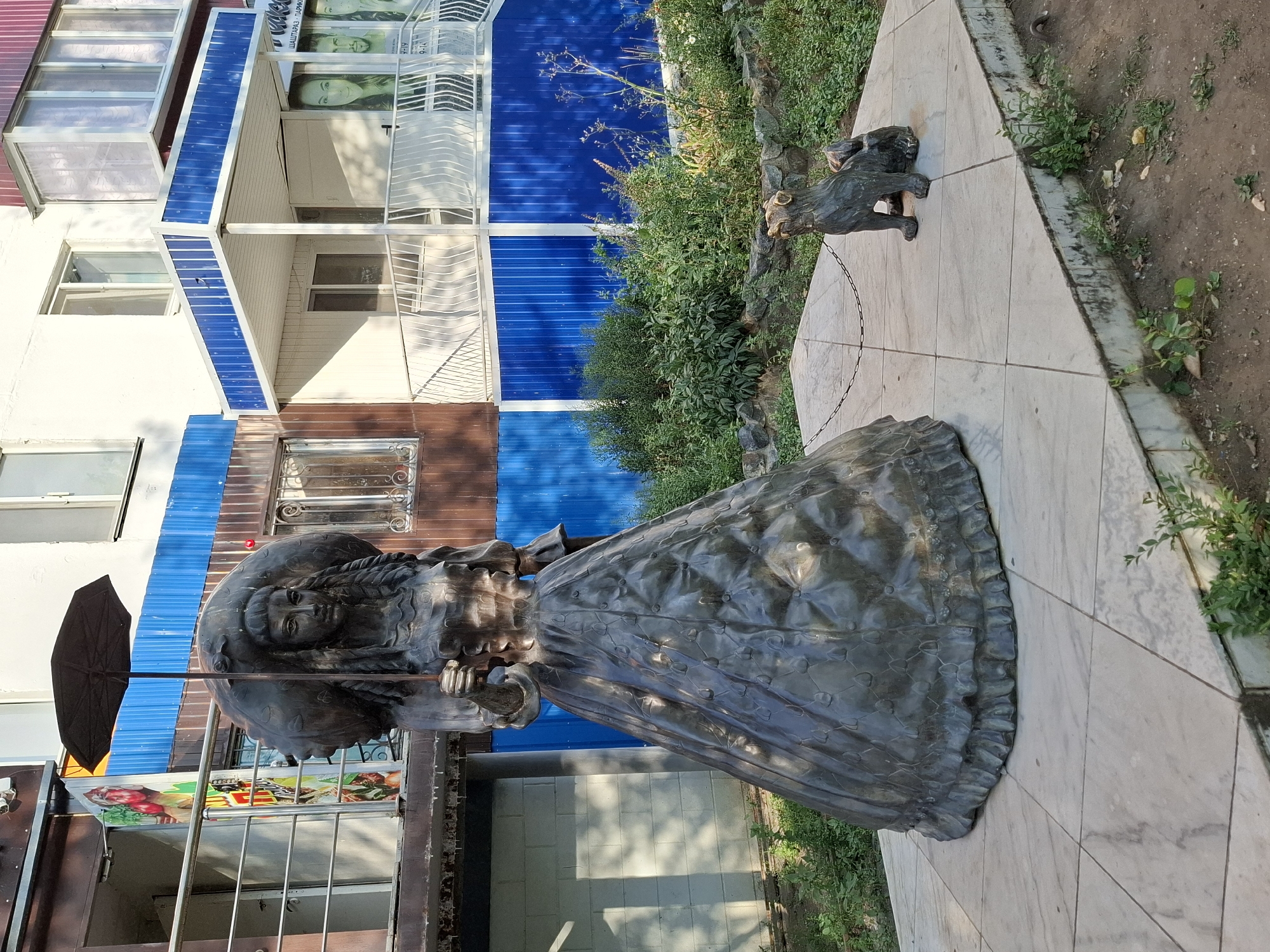 This image shows a bronze sculpture of a woman holding an umbrella and a large bundle, standing on a tiled platform. To the right of the woman, a smaller bronze sculpture of a dog on a chain is visible. The sculptures are located in an outdoor setting, with a building in the background featuring blue and brown siding, balconies, and windows. There is greenery and vegetation around the platform. This sculpture is located in the city of Lisakovsk, Kazakhstan, specifically in front of the building at 19 Mir Avenue.