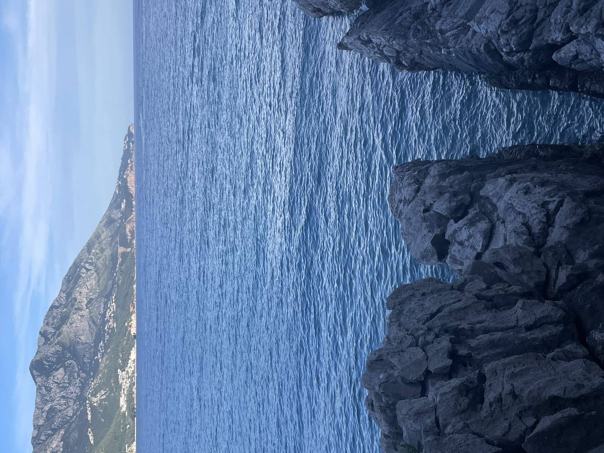 The image depicts a scenic coastal view with a mountainous landscape in the background and rocky cliffs in the foreground. The sea occupies a large portion of the frame, with gentle ripples on its surface. In the distance, a mountain range rises, dotted with buildings that suggest a settlement or town. The sky above is a pale blue with scattered clouds. The foreground is dominated by dark, jagged rocks that form a natural barrier or overlook to the sea. The scene appears to be a natural landscape, likely a coastline, with no human figures or explicit signs of human activity beyond the distant buildings. The lighting suggests it is daytime, possibly late afternoon or early morning given the soft light on the mountains. This view is characteristic of the coastal areas near Budva, Montenegro, known for its mountainous terrain meeting the Adriatic Sea and picturesque towns.