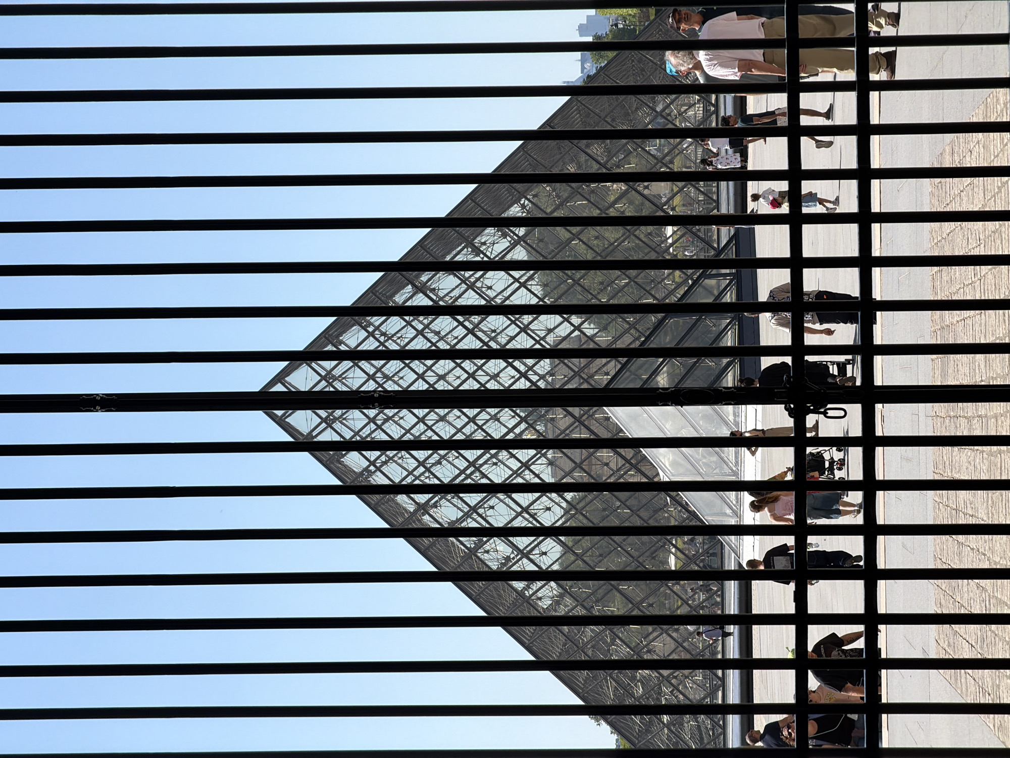 The Louvre Pyramid is viewed through open Venetian blinds, with the slats oriented horizontally. People, indistinct and small, are seen walking outside the pyramid. The sky is pale blue. The perspective is from inside a building. This image was taken in Paris, France.