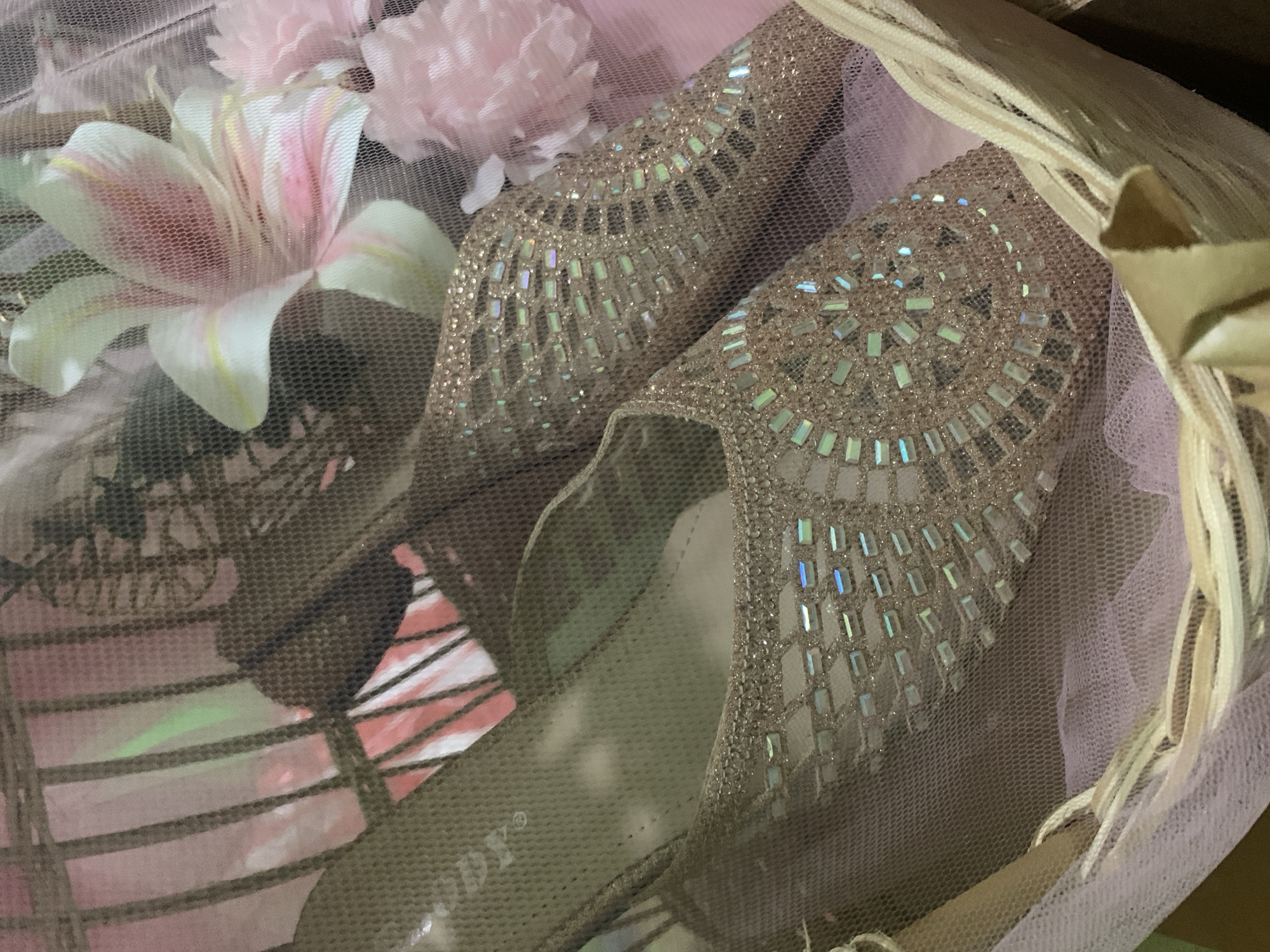 The image shows a pair of sparkly, closed-toe shoes with intricate embellishments, resting in a basket. The shoes are a light, shimmery color, possibly rose gold or champagne, and are adorned with small, rectangular jewels that catch the light and create a holographic effect. The basket itself appears to be made of woven material and is lined with sheer, pink fabric, suggesting a decorative presentation. Behind the shoes, a blurred background reveals elements of what looks like decorative flowers and perhaps some other packaging material. The overall impression is one of a gift or a special occasion item. There are no people or identifiable landmarks visible in the image. The location in Kuala Pembuang, Indonesia, is not discernible from the visual information presented.