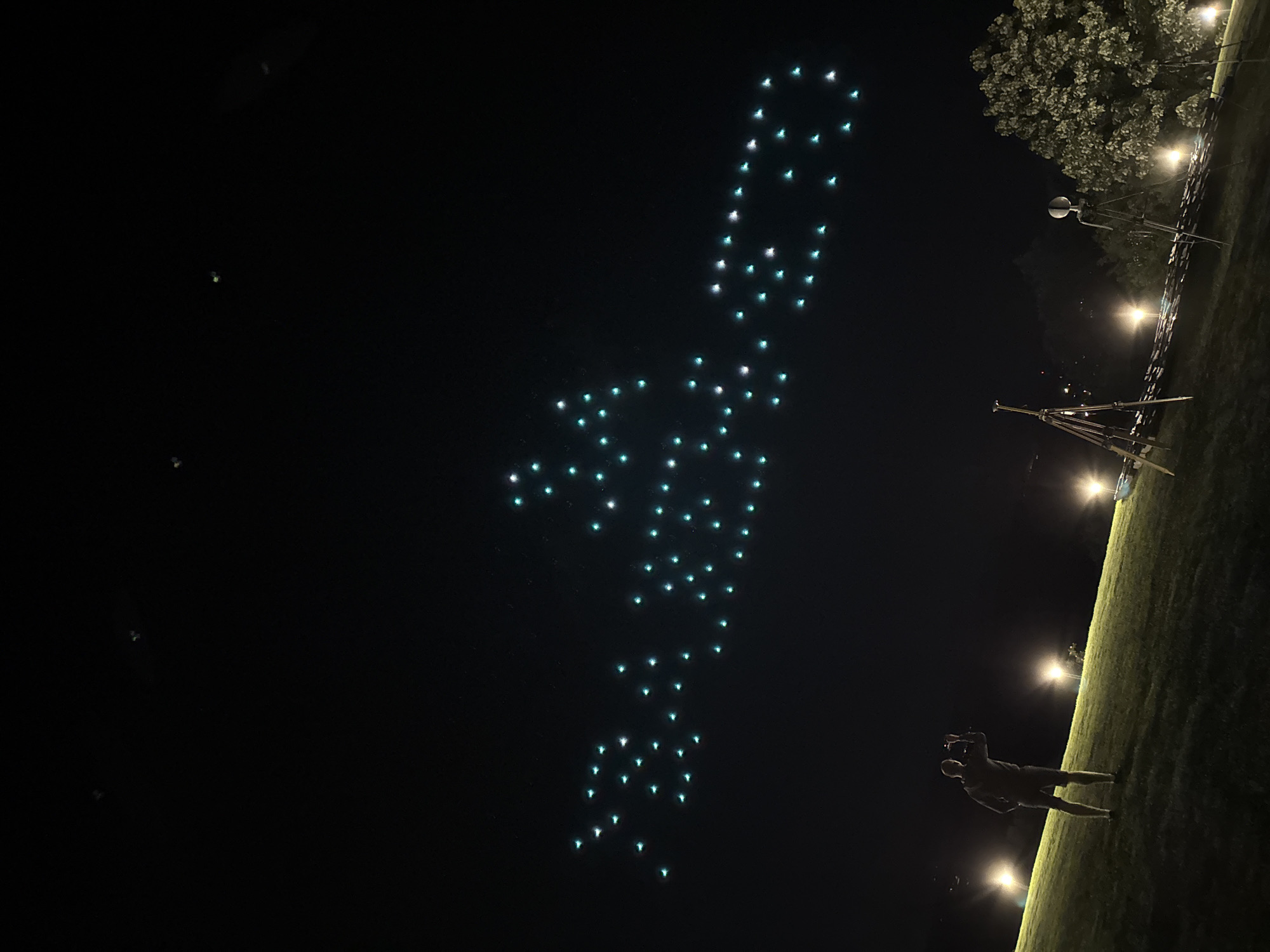 In Baliovtsi, Bulgaria, at night, a drone show displays the word "BALIO" in the sky, formed by numerous small, bright lights, mostly teal and white. The lights are arranged in a pattern that spells out the word. In the foreground, a grassy slope is illuminated by several streetlights. A man stands on the slope, facing away from the viewer, holding what appears to be a camera or phone, capturing the drone display. To the right, two tripods are set up, one with a device on top, and a skeletal figure-like object. A large tree with foliage is visible on the right side of the frame. The sky is dark, indicating nighttime.