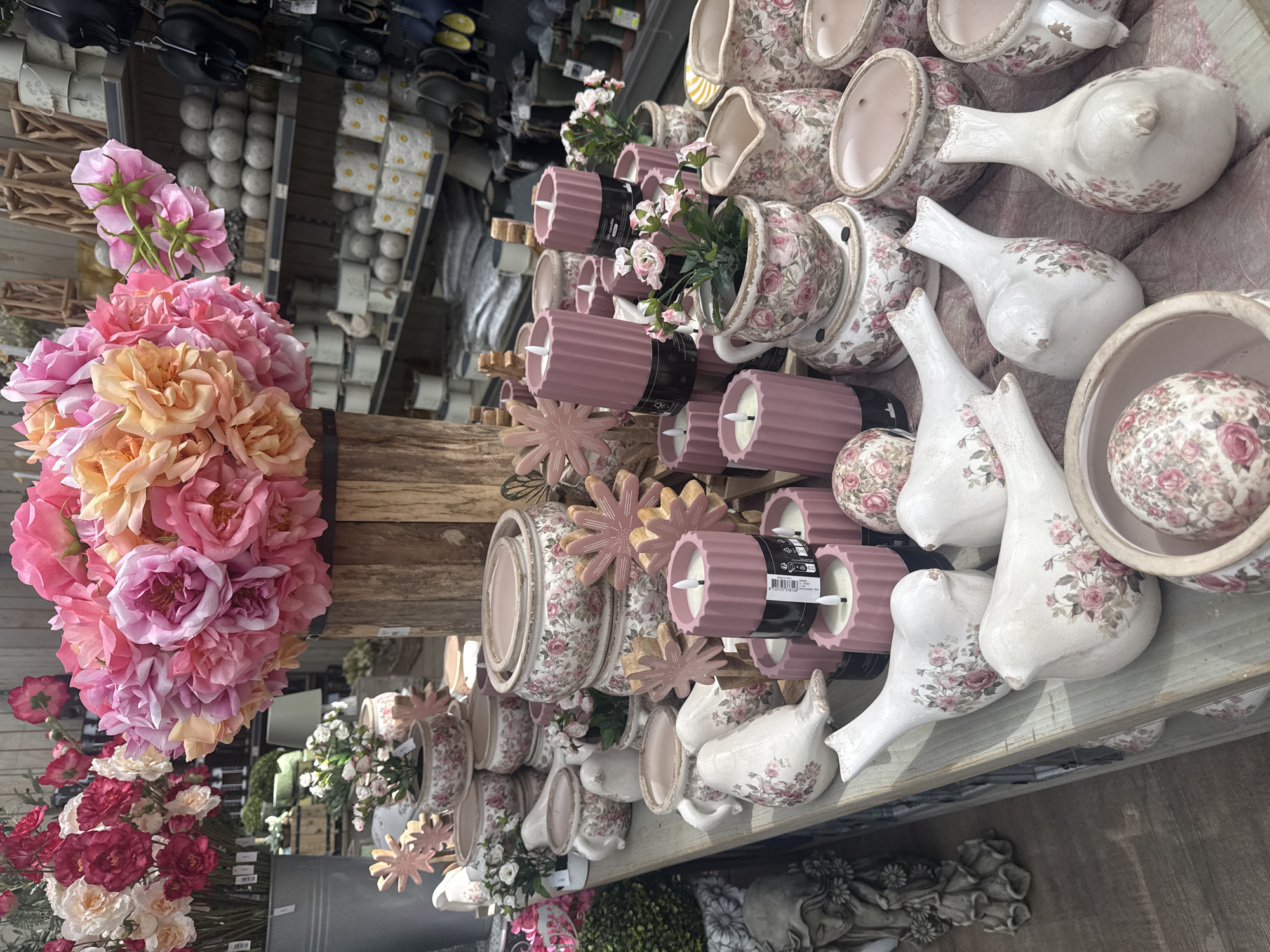 In Fritzlar, Germany, a retail display features a collection of floral-themed decorative items. The foreground is dominated by several ceramic bird figurines, each adorned with pink and white floral patterns. Adjacent to the birds are cylindrical pink candles with visible wicks, some stacked and others individually placed. Various ceramic pots and planters, also decorated with similar floral motifs, are arranged throughout the display. A large bouquet of artificial pink and orange roses is prominently featured in a wooden barrel towards the top of the frame. In the background, shelves display more ceramic items and other merchandise, suggesting an indoor retail environment. The overall impression is one of a curated display of decorative home goods, likely in a garden center or gift shop.